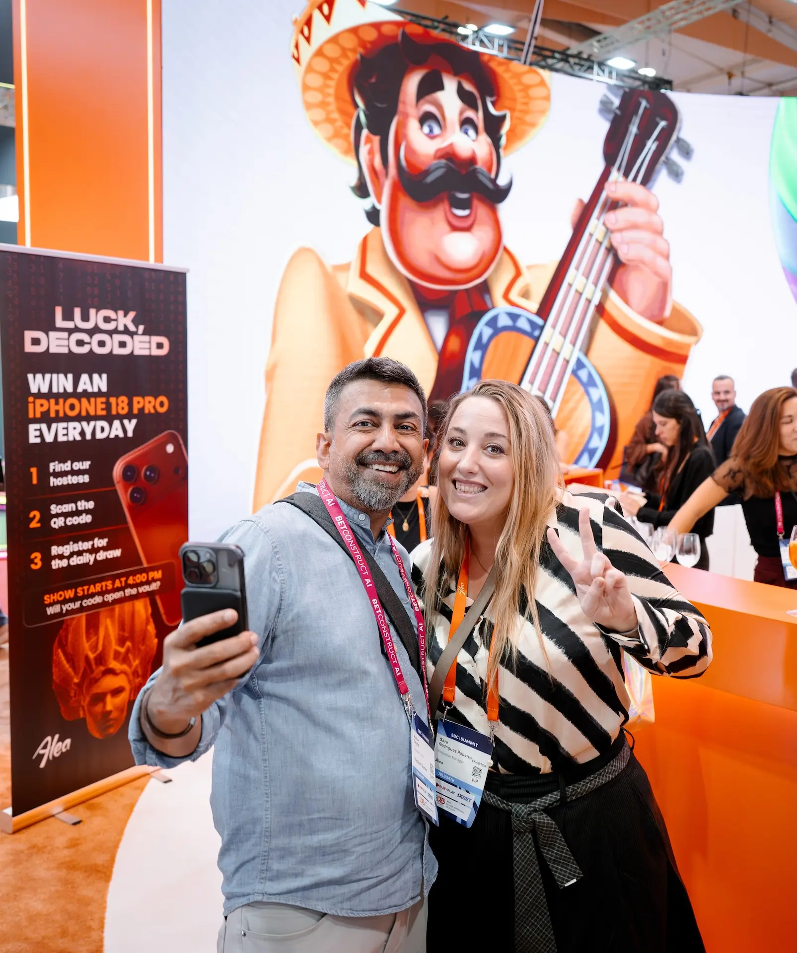 Two visitors taking a selfie at the Alea stand in front of the mariachi mascot and the iPhone 18 Pro raffle banner, SBC Summit Lisbon 2026