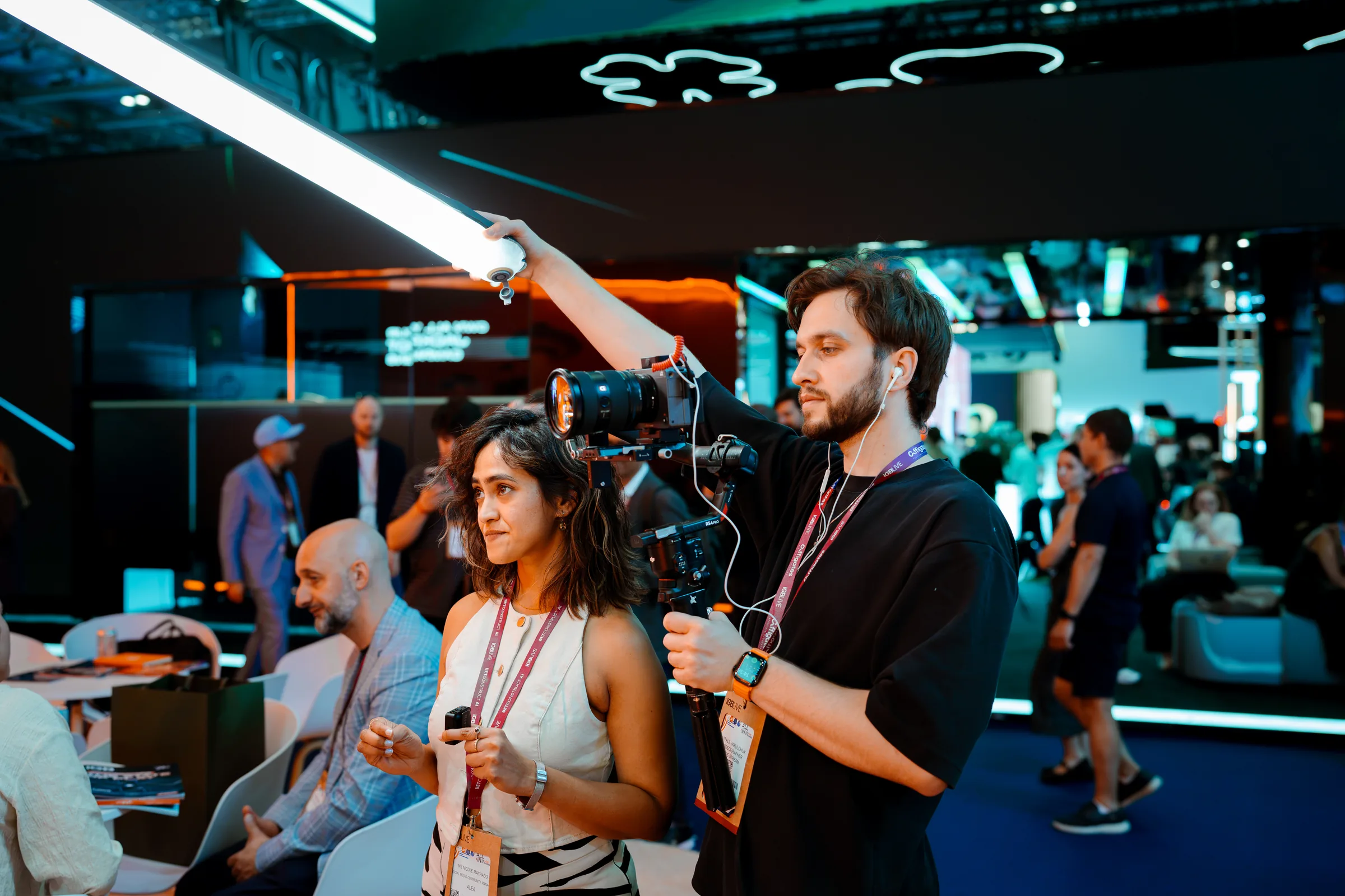 An operator filming with a camera in one hand and a light in the other while a client talks to the stand team