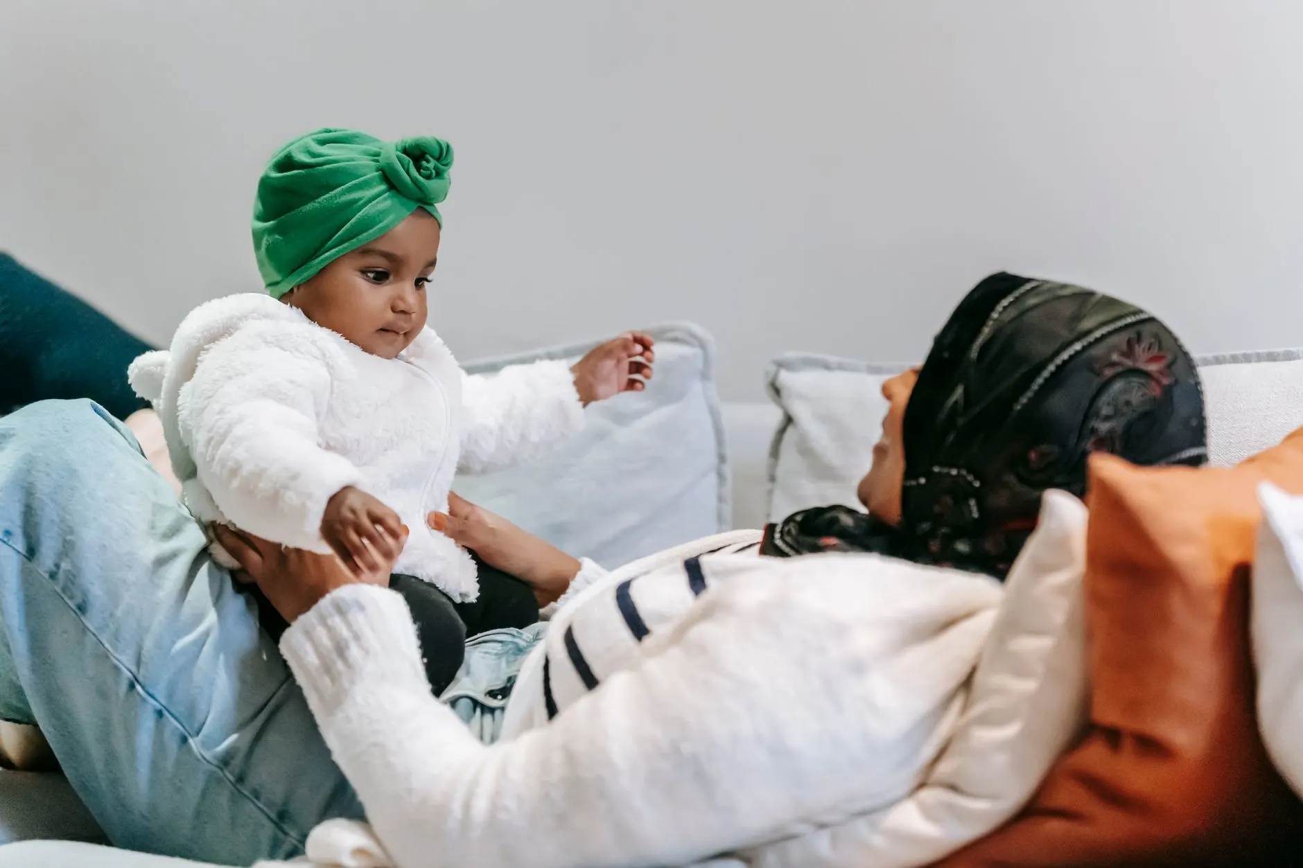 Cheerful African American mom in hijab lying on sofa with cute little daughter wearing turban