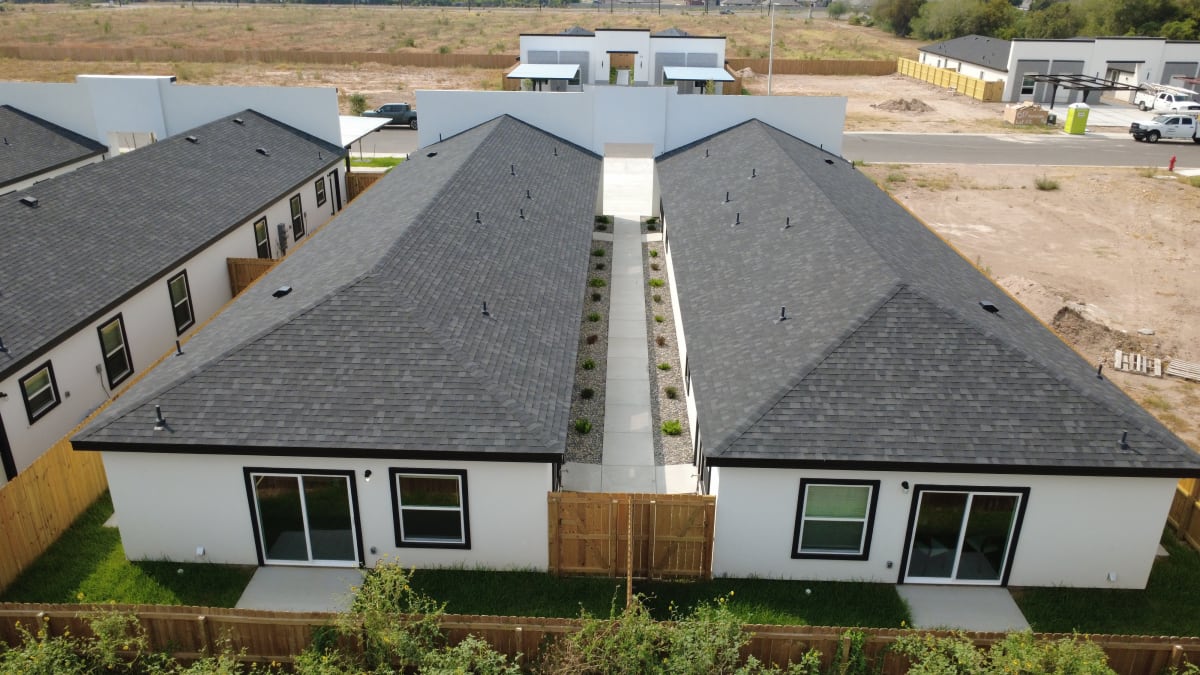 Rear of a fourplex from above: two hipped-roof wings, a planted breezeway, sliding patio doors and wood-fenced yards