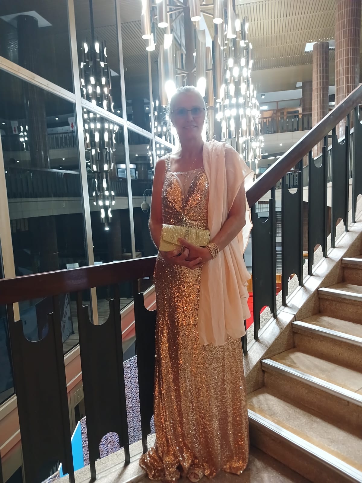Magriet .jpeg A woman standing on a staircase. she is wearing a beautiful silver evening dress and holding a clutch purse.