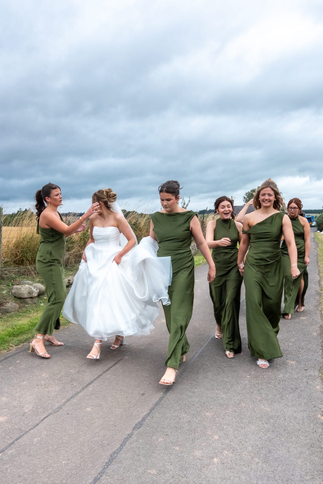 The bridesmaids in green walk with Ellie beneath a cloudy sky