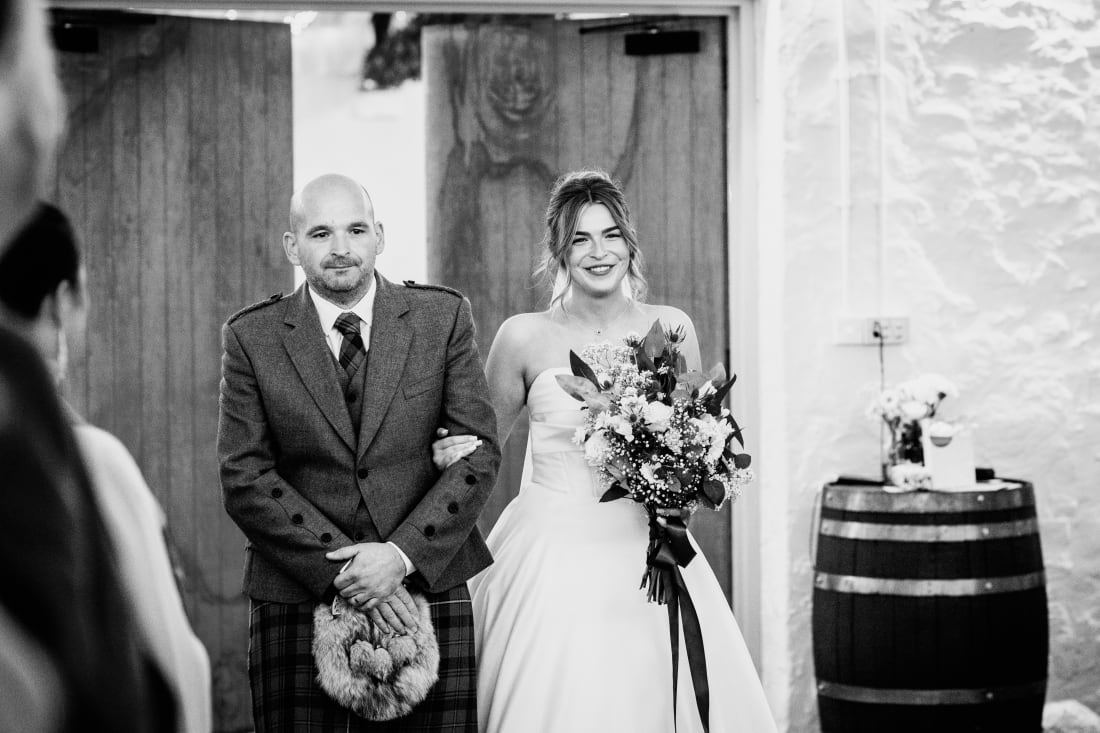 Ellie walks down the aisle on the arm of a guest in a kilt