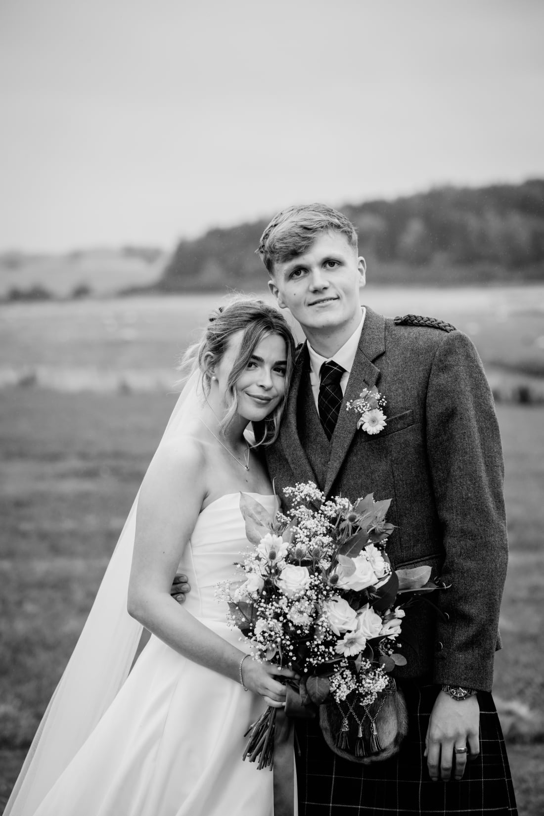 The couple stand together in the fields holding the bouquet