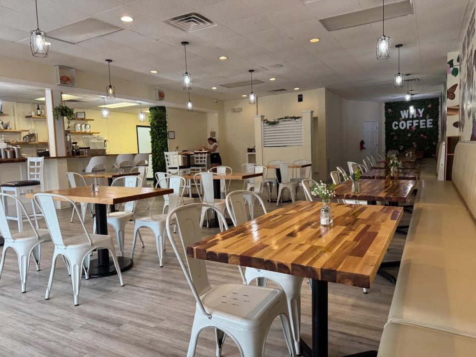 Bright dining room with wooden tables, white metal chairs, pendant lights, and a green wall reading WHY COFFEE.