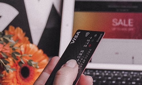 Climate change action - reduce consumption. Image shows a closeup of a man's hand holding a black debit card in front of a laptop screenedit card