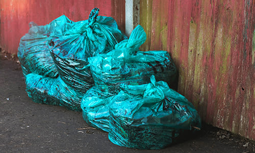 Climate change action - reduce waste. Image shows a pile of bright blue rubbish bags (garbage bags) in front of a tatty red painted fence