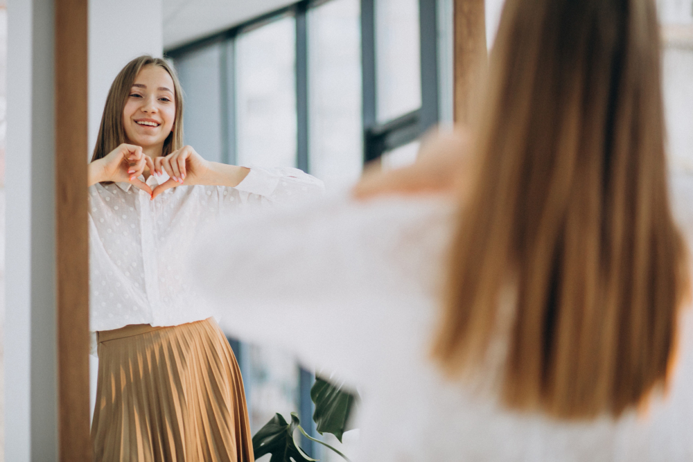 Jeune femme qui se regarde dans le miroir et qui forme un cœur avec ses mains en signe d’amour de soi