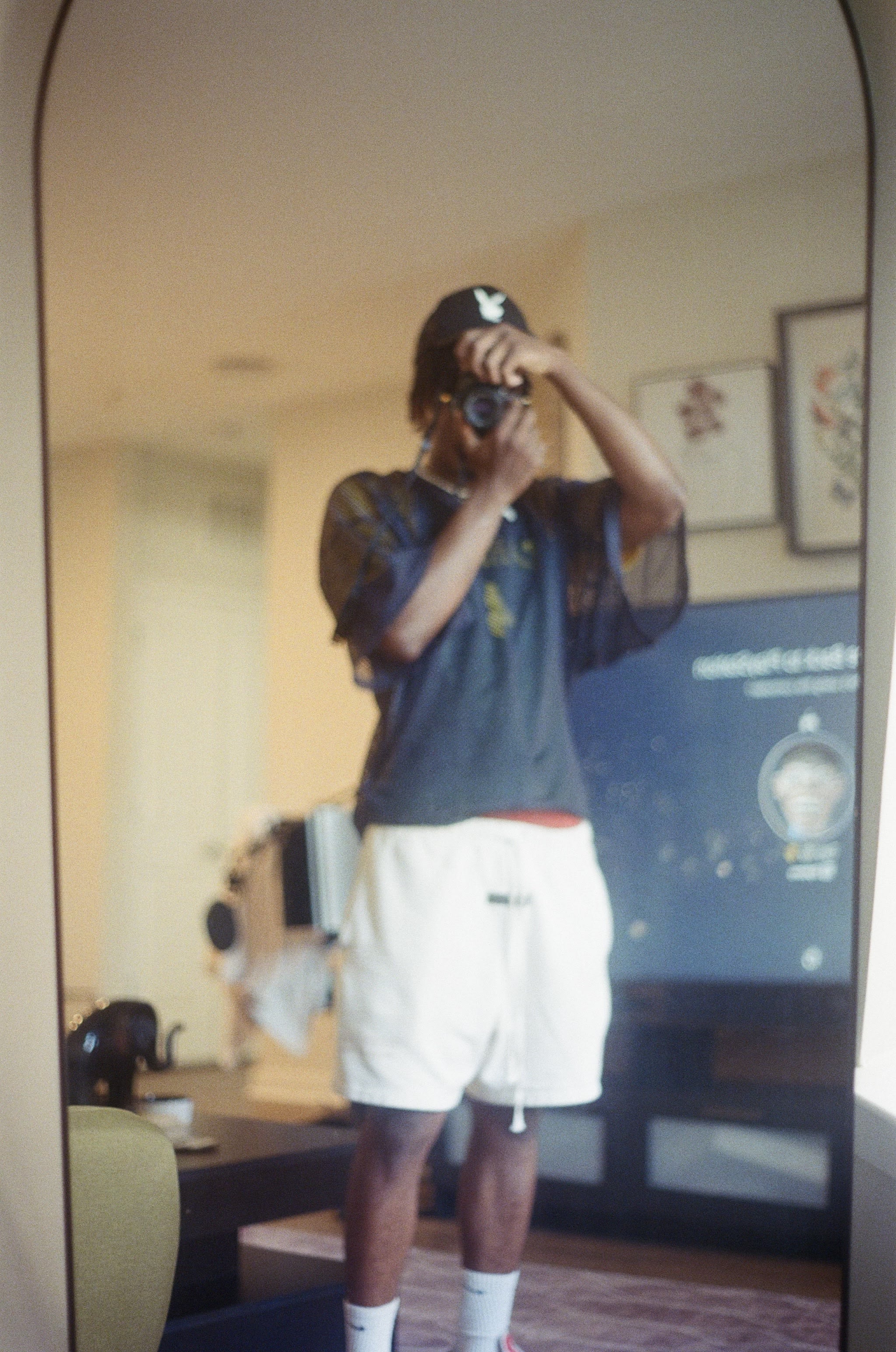 Mirror selfie of Kama in a Playboy trucker cap and mesh tee, film camera up to his eye