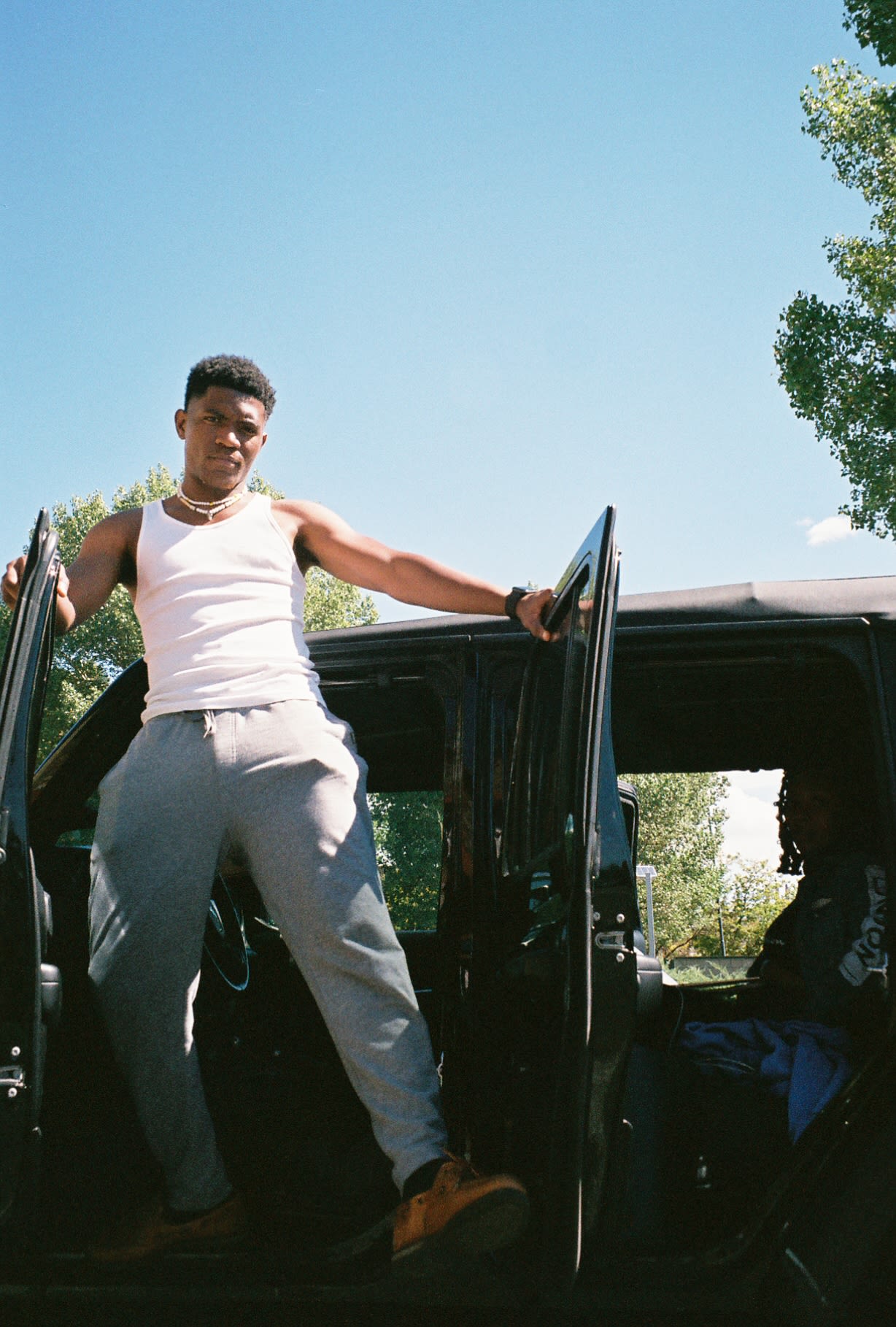 Shot on film: Kama in a white tank standing tall in the door of a black Jeep, friend in the back seat