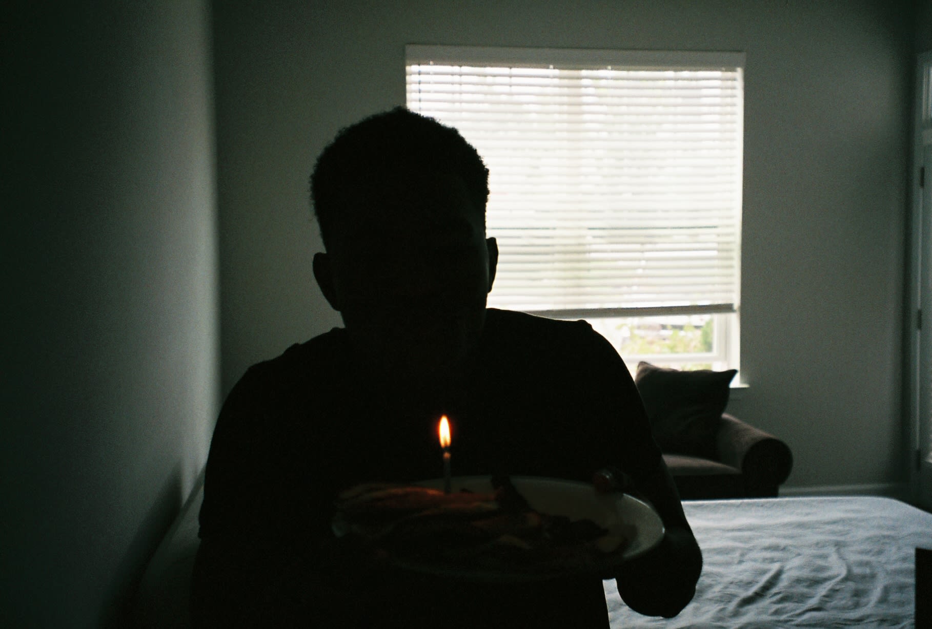Shot on film: silhouette of Kama holding a plate with a single lit birthday candle by the window
