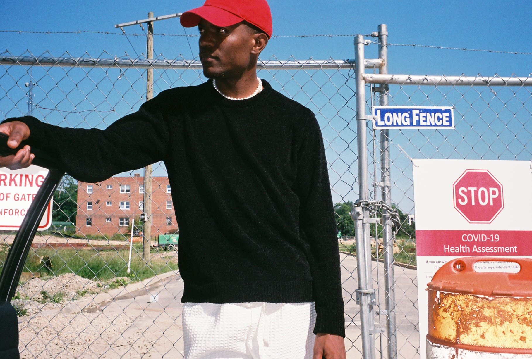 Shot on film: Kama in a red cap, black sweater, and pearls leaning on a car door by a chain-link gate
