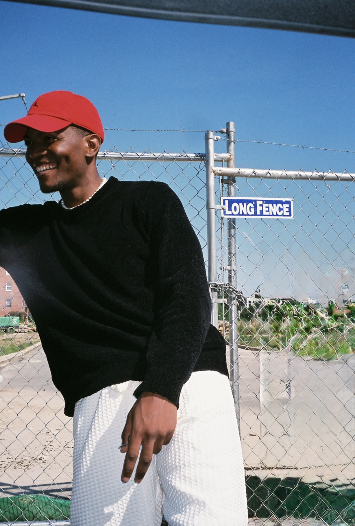 Shot on film: Kama laughing in a red cap and black sweater against a chain-link fence and blue sky