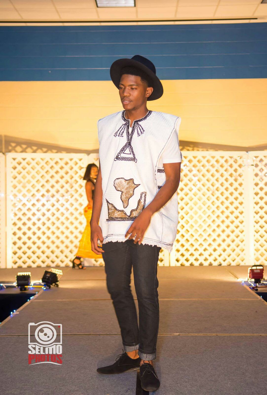Throwback: teenage Kama walking a runway in a wide-brim hat and white tunic with an Africa applique