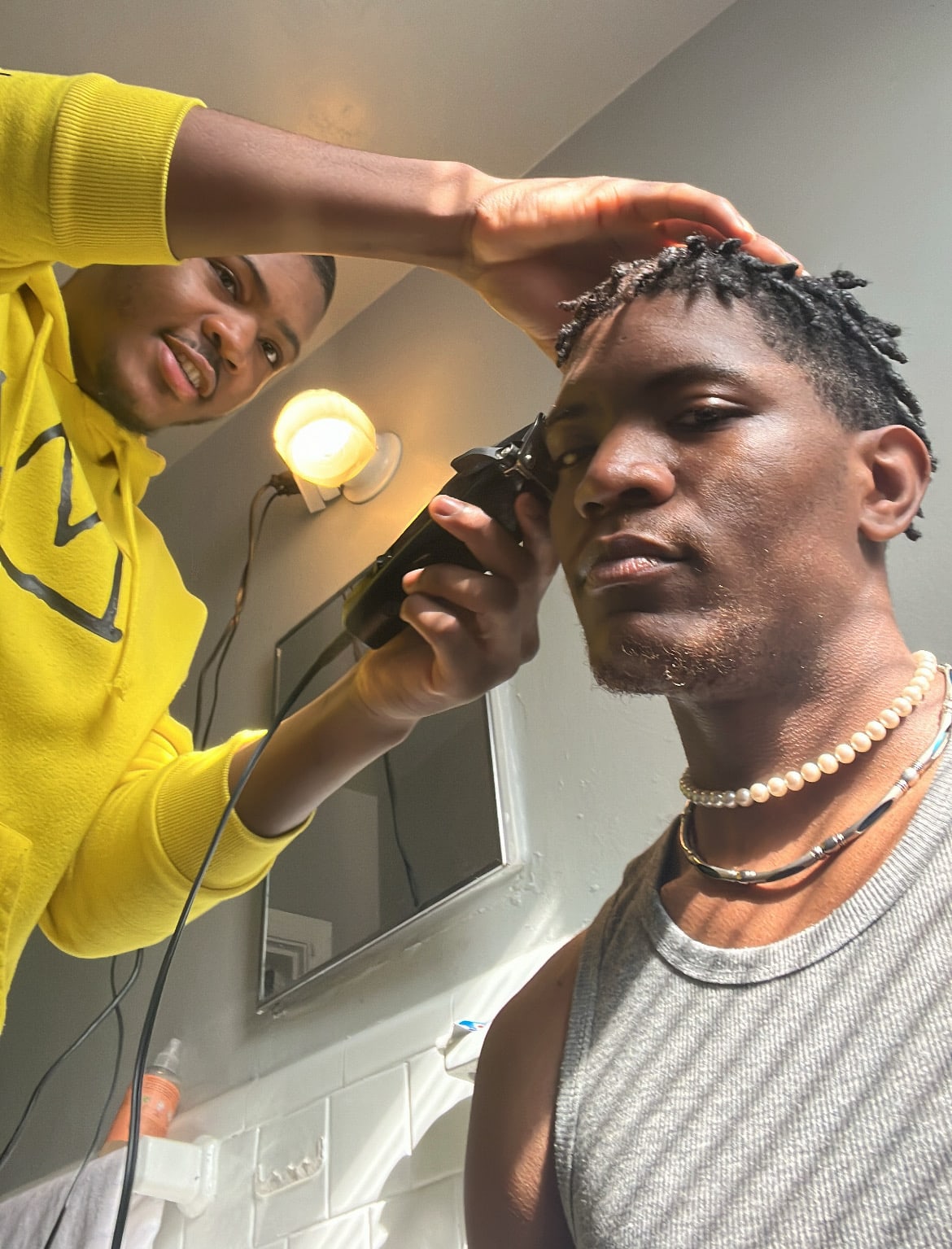 A friend in a yellow hoodie lining Kama up with clippers in the bathroom mirror light