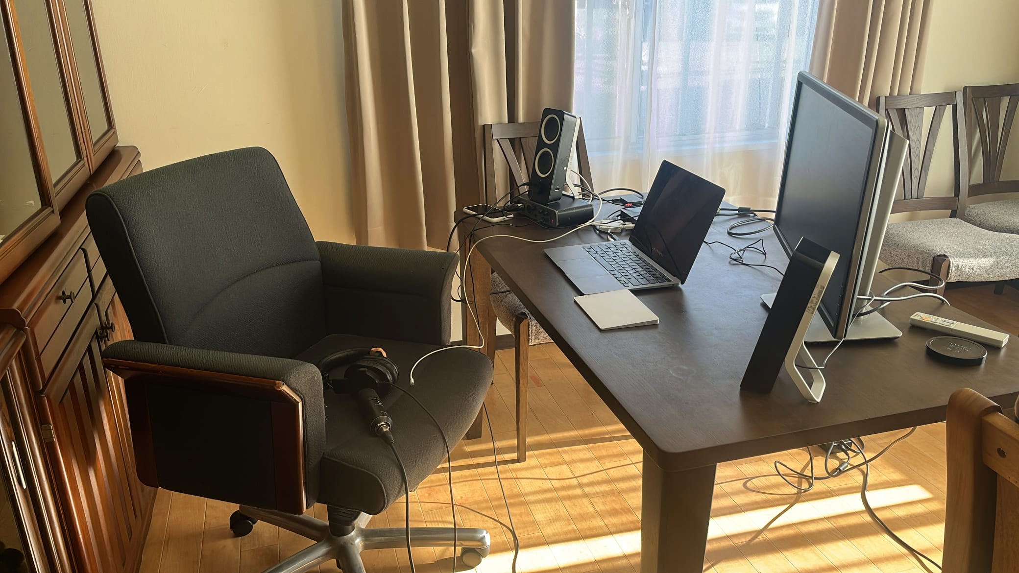 A sunlit desk setup - laptop, monitor, speakers, and headphones resting on the chair