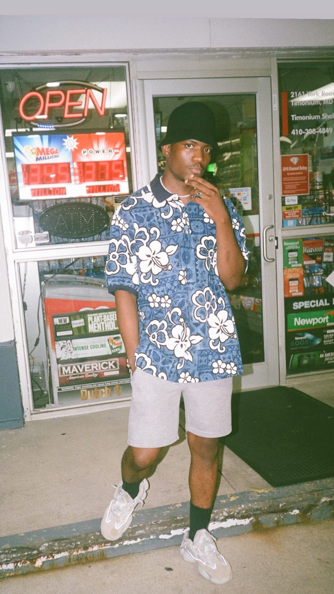 Kama in a blue floral shirt and bucket hat outside a corner store at night, OPEN sign glowing