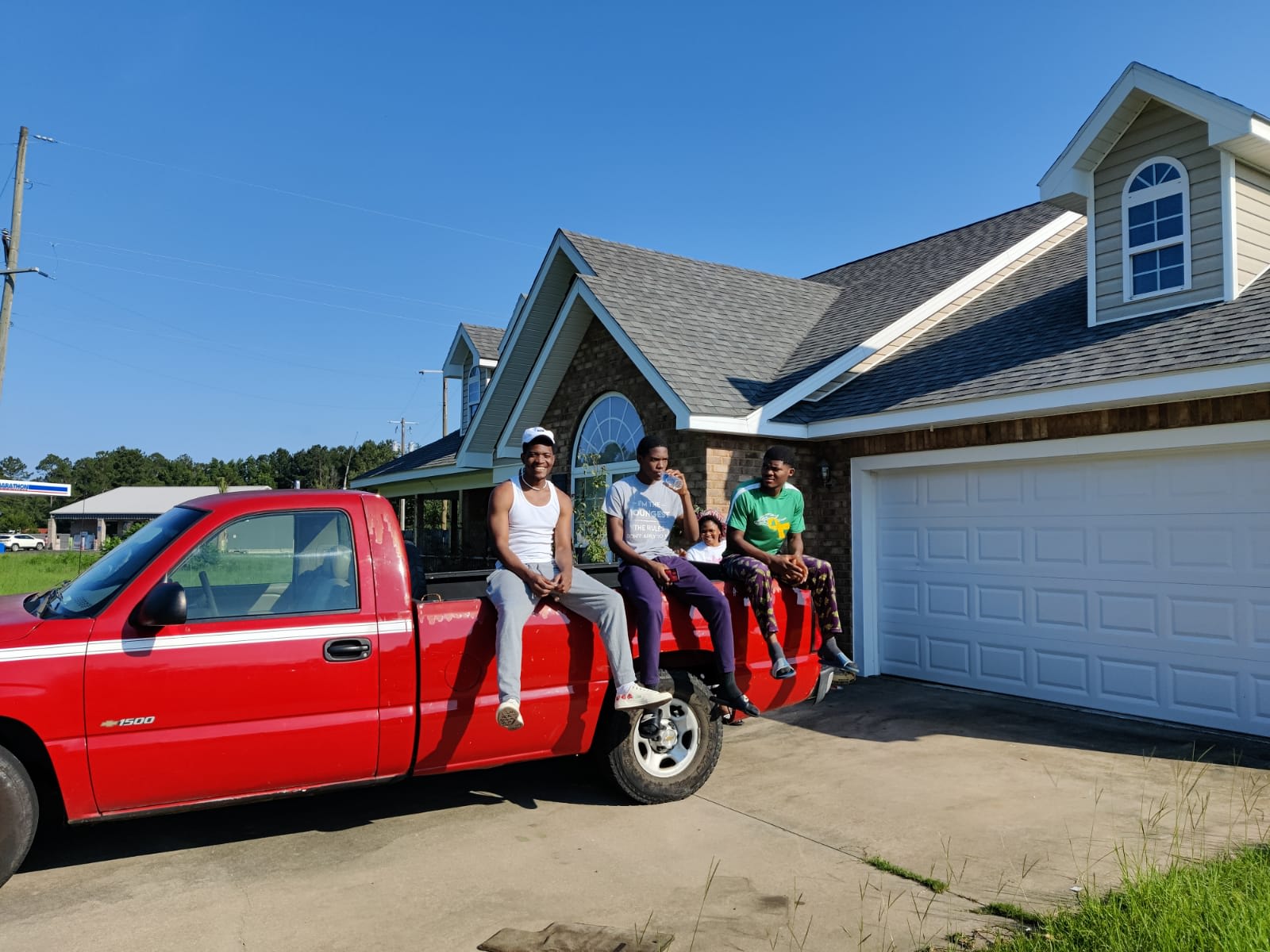 Four friends perched on the bed of a red pickup outside the house, all blue sky