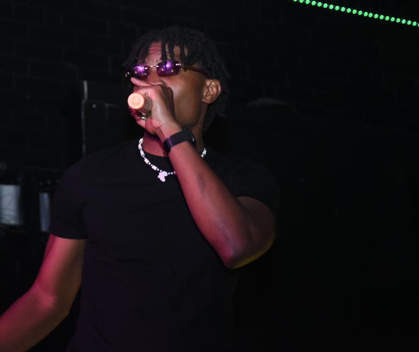 Kama in shades on the mic under green light, mid-performance on a dark stage