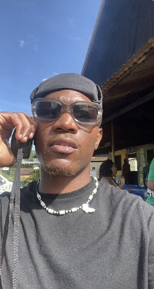 Sunny selfie of Kama in tinted glasses and a beaded Africa-pendant necklace at an open-air roadside spot
