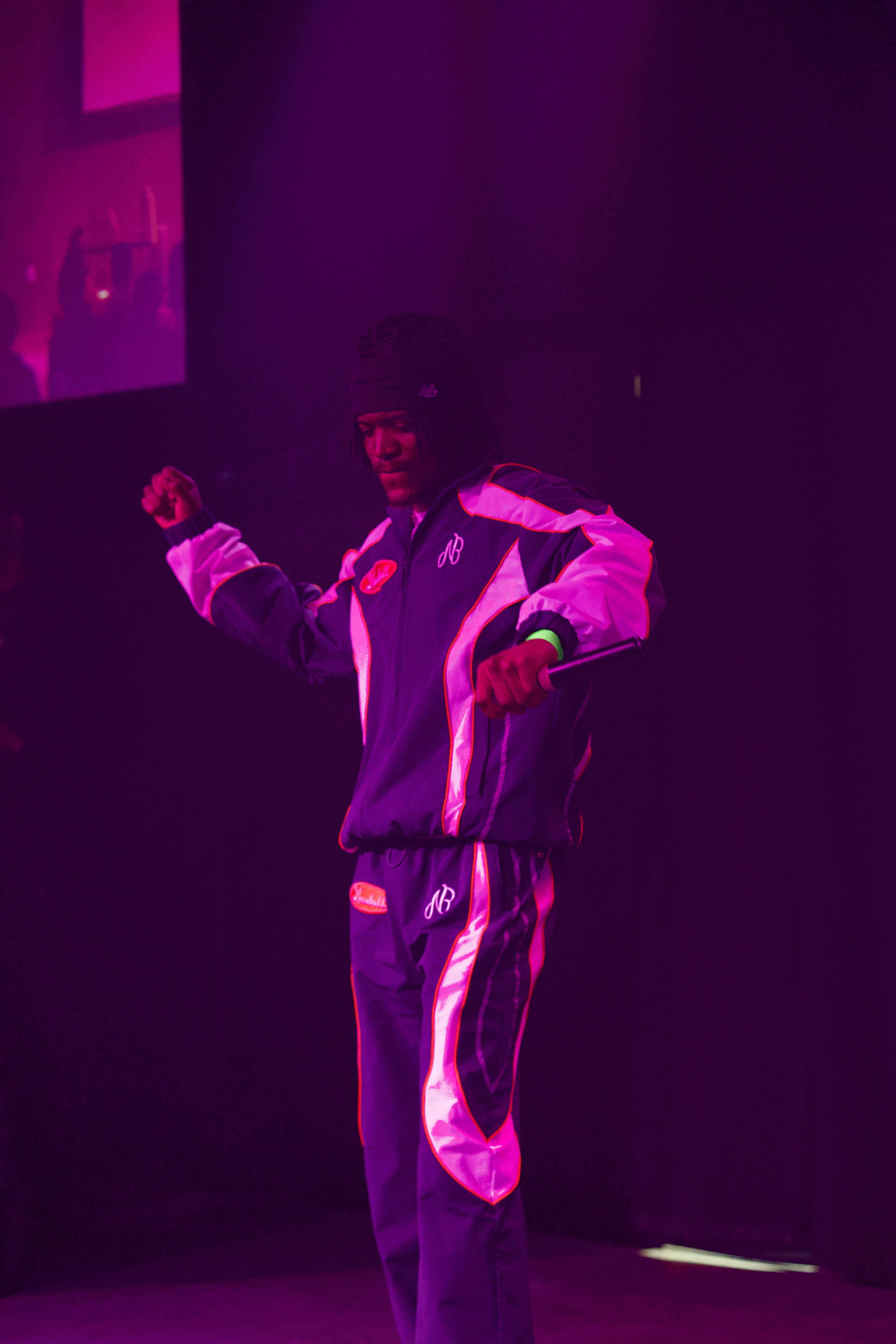 Kama dancing on stage in a racing tracksuit, fist up, bathed in pink light