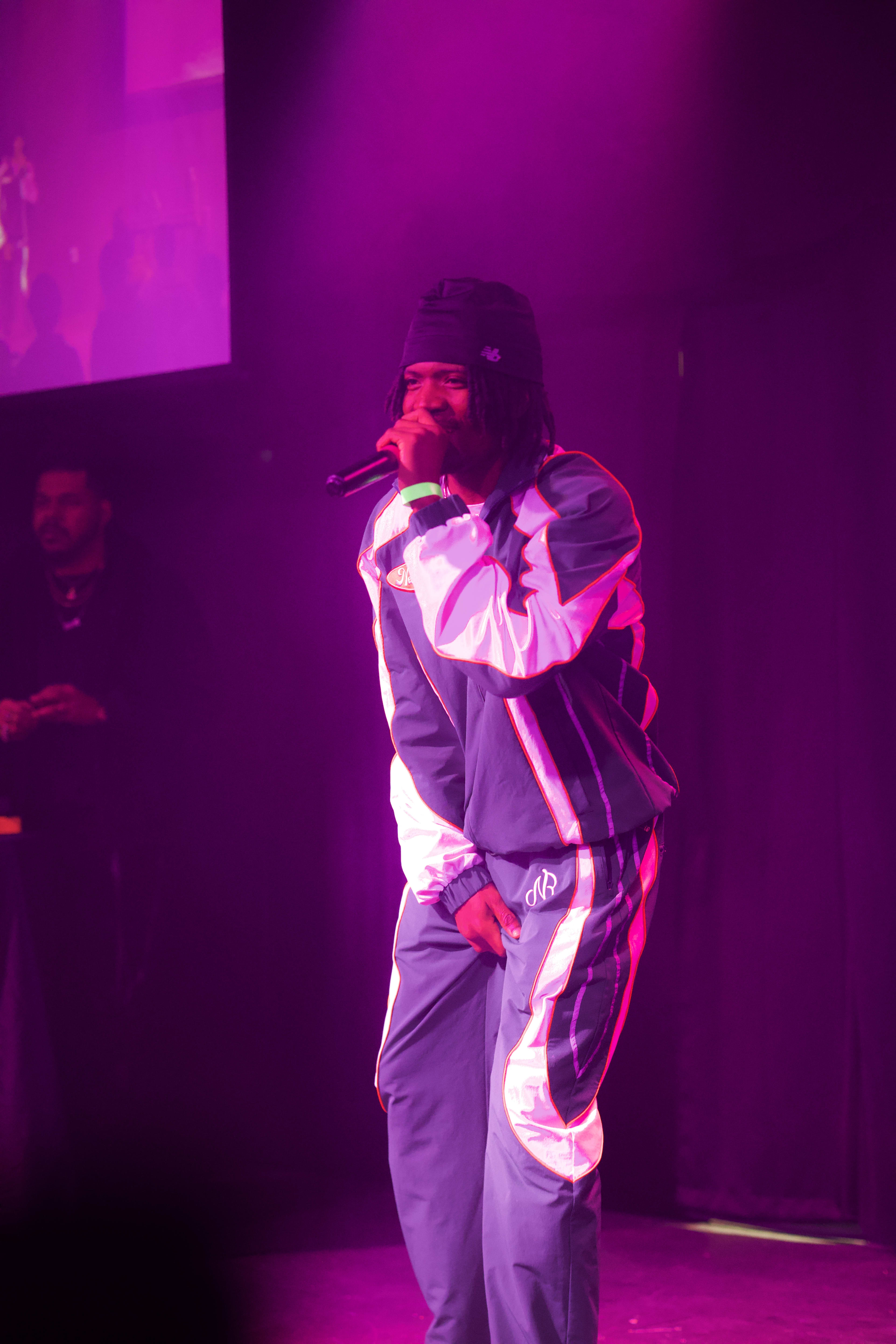 Kama singing into the mic on stage, tracksuit catching the purple light