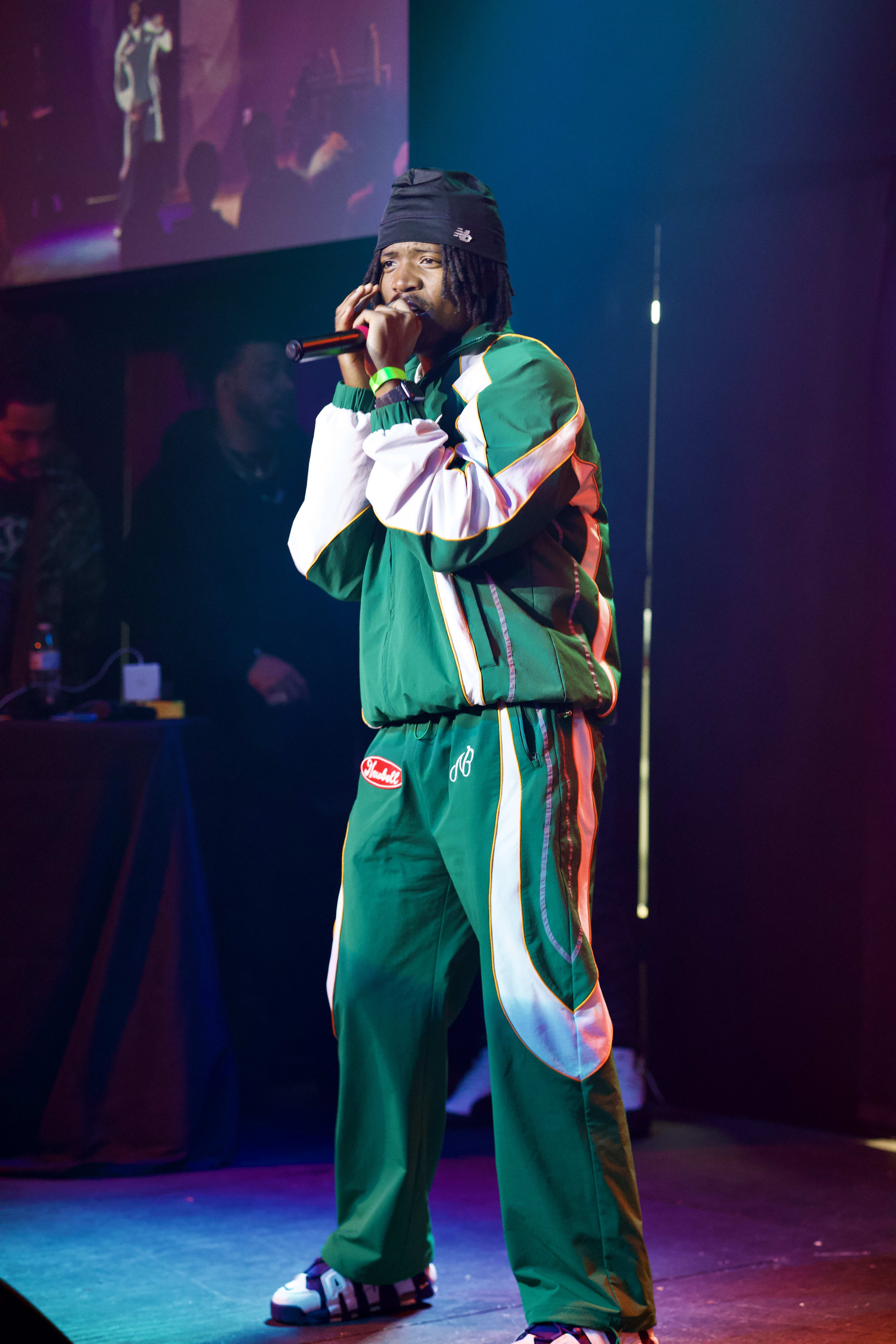 Kama on stage in a green-and-white tracksuit, both hands on the mic