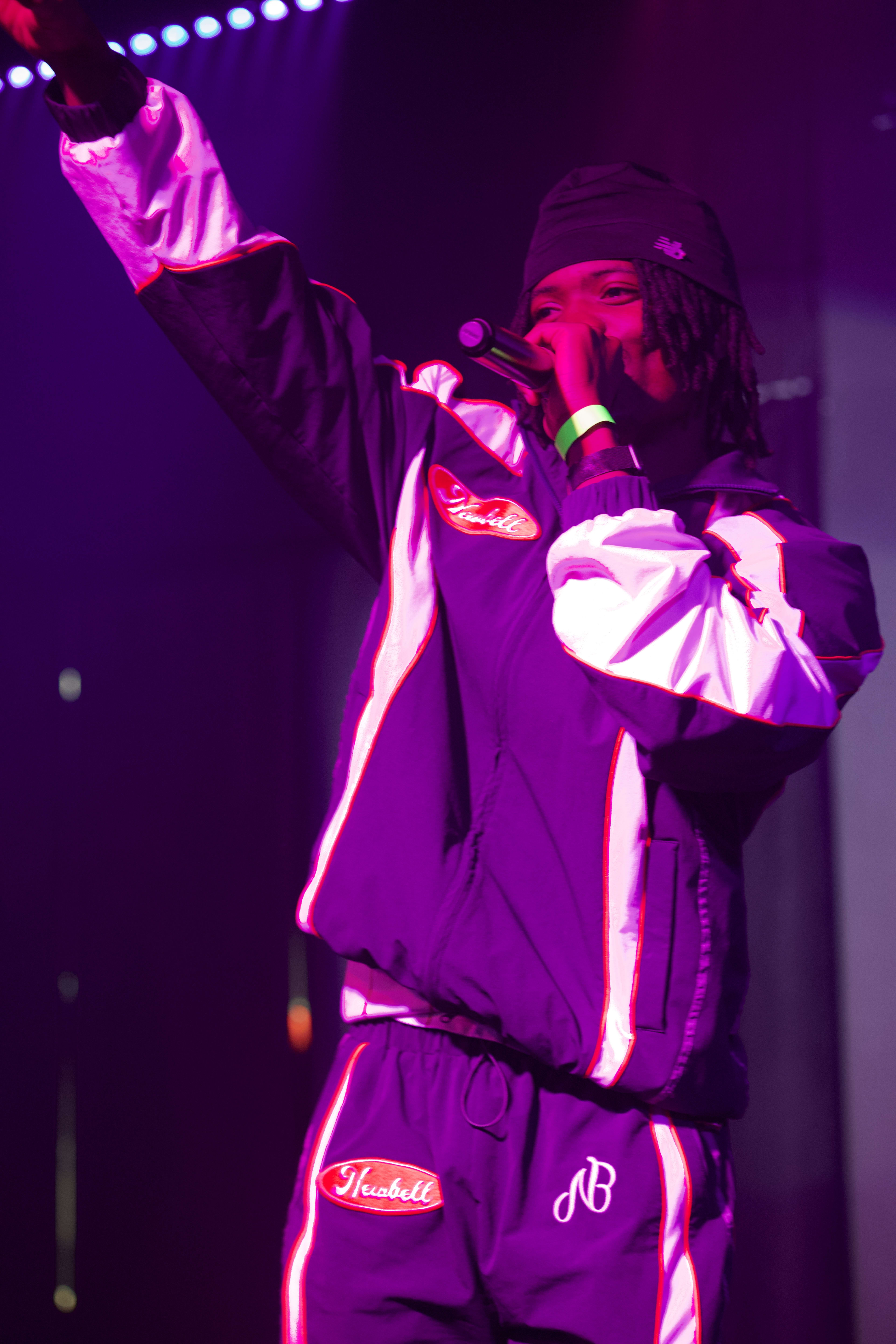 Kama mid-performance, one arm raised high, tracksuit glowing under the stage lights