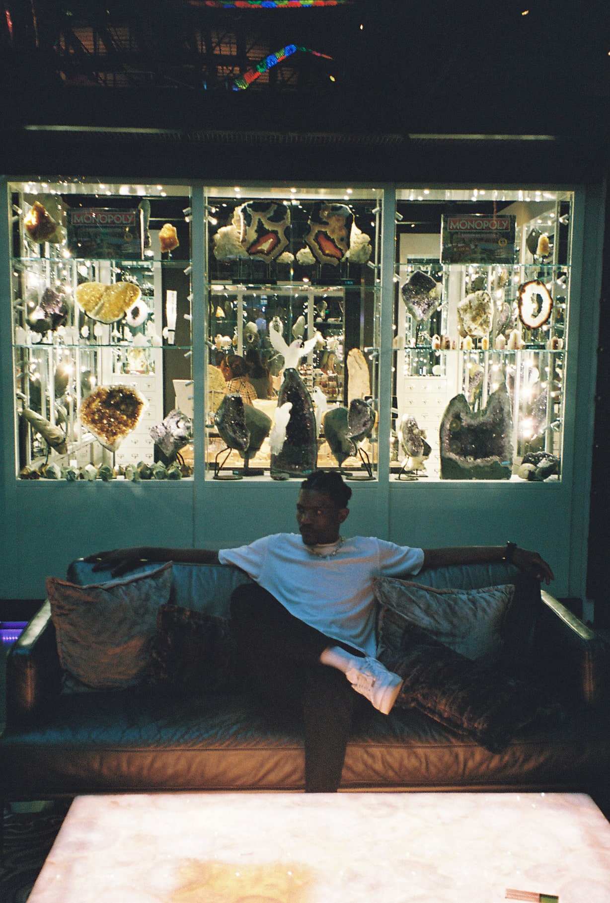 Shot on film: Kama stretched arms-wide on a couch in front of a glowing crystal display case