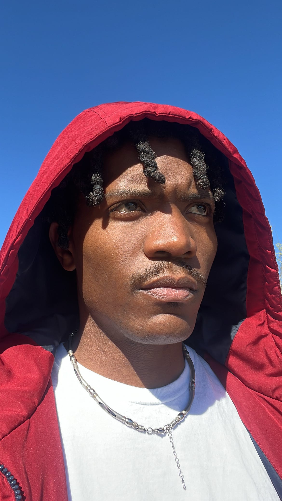 Close selfie of Kama in a red hood and chain against a cloudless blue sky