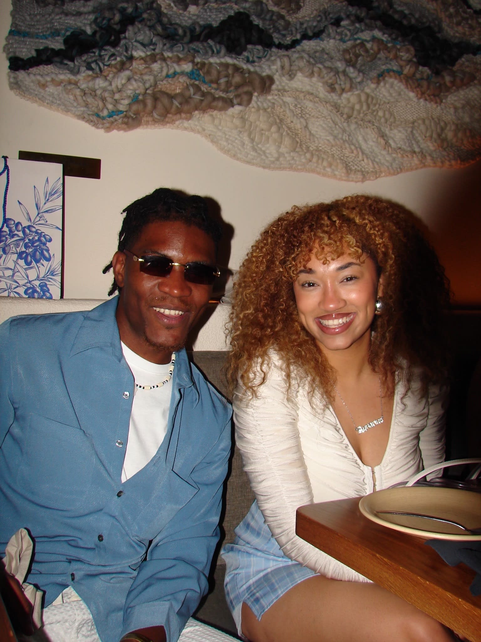 Kama in a light-blue shirt and sunglasses beside a friend with golden curls at a restaurant booth