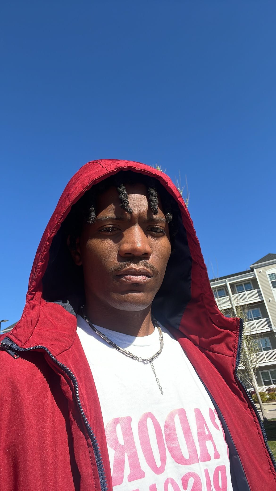 Sunny selfie of Kama in a red hooded jacket and silver chain, apartment balconies behind