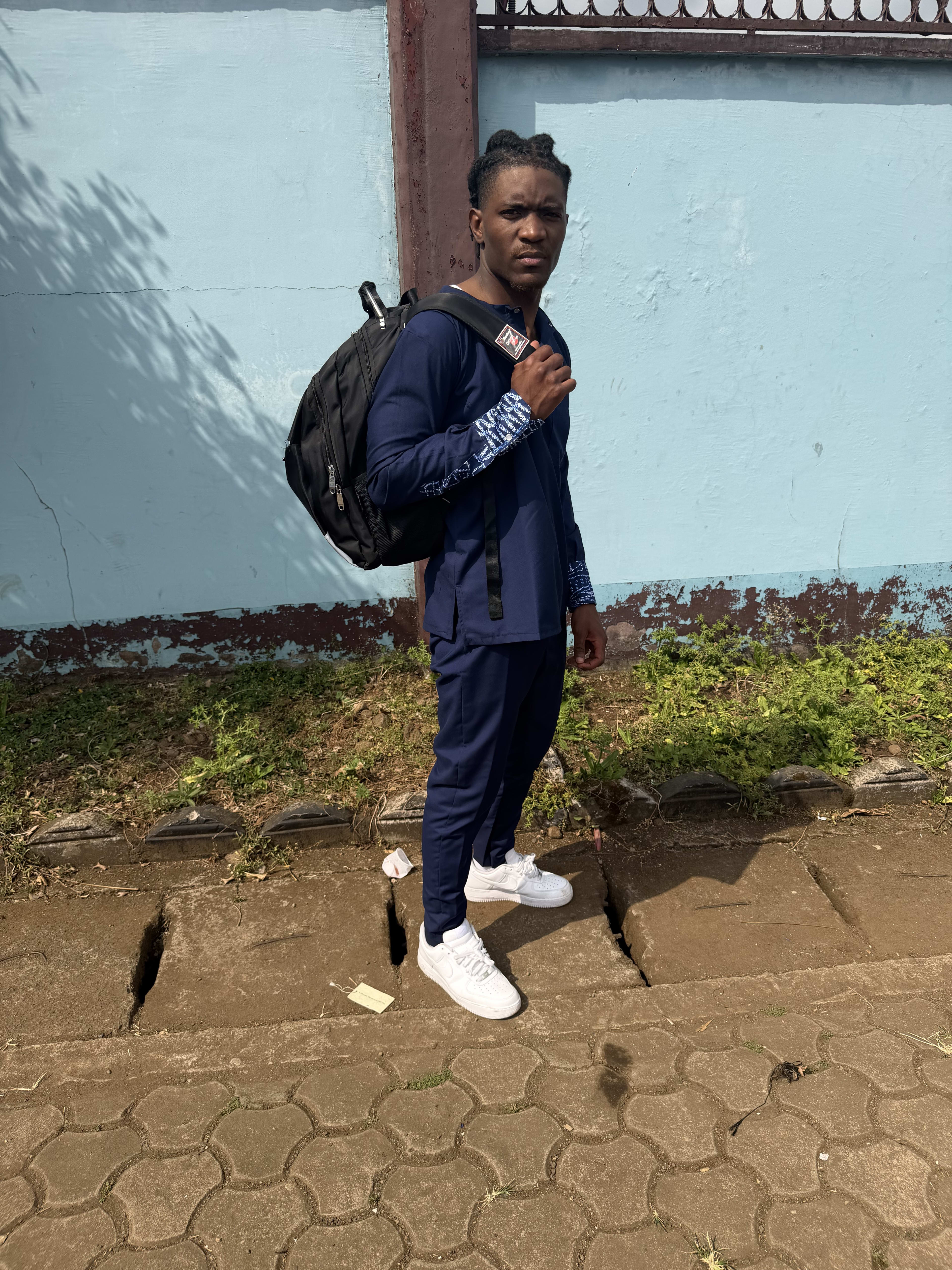 Kama with a backpack slung on one shoulder, standing on a cracked-brick walkway by a blue wall