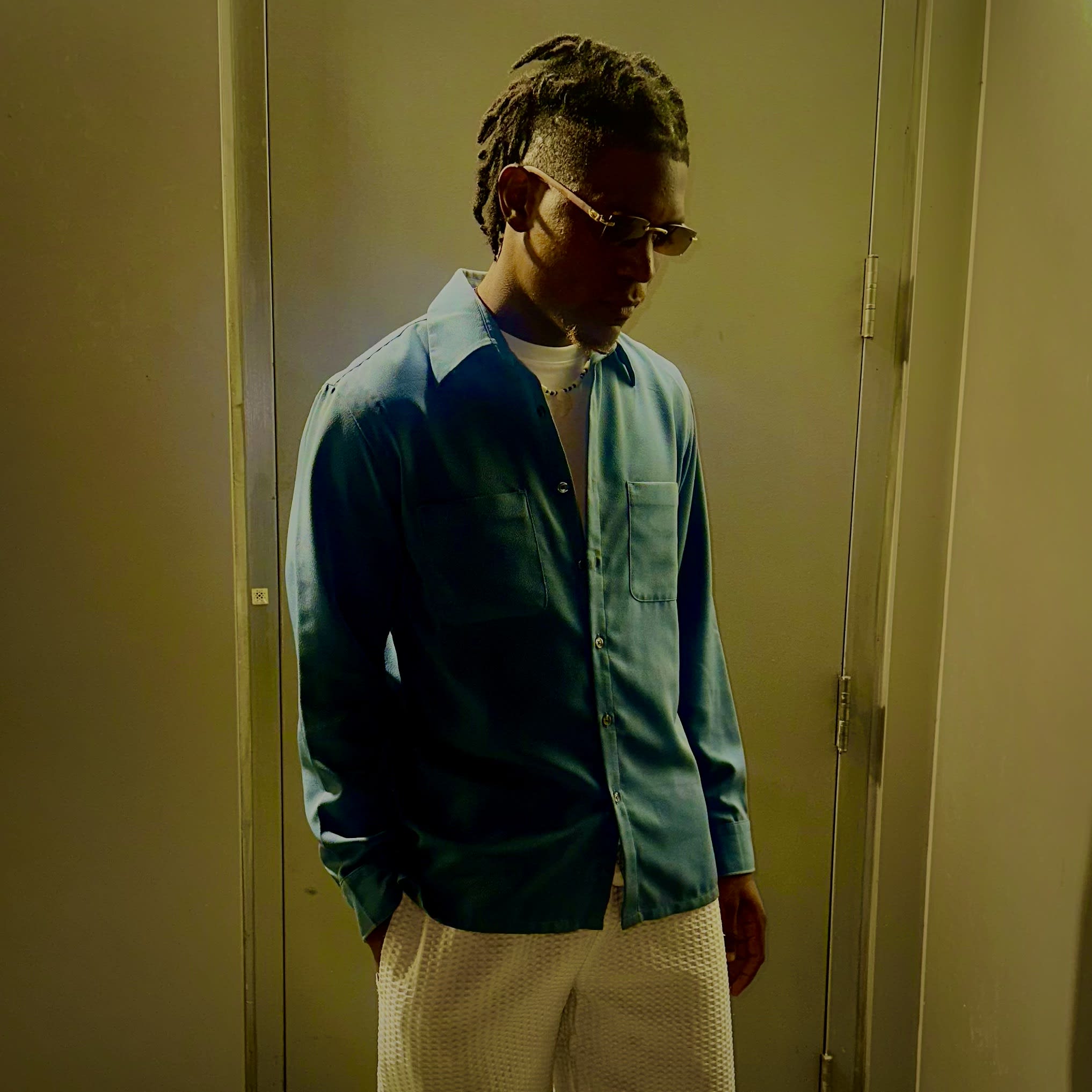 Kama in a light-blue button-up and white mesh shorts, head bowed under the hallway light