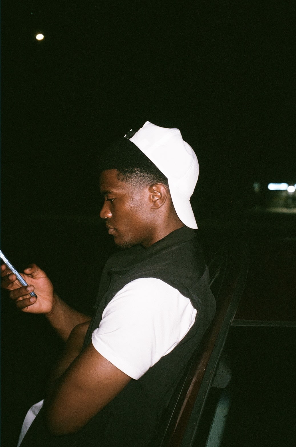 Shot on film: Kama in a backwards white cap checking his phone against a car in the dark