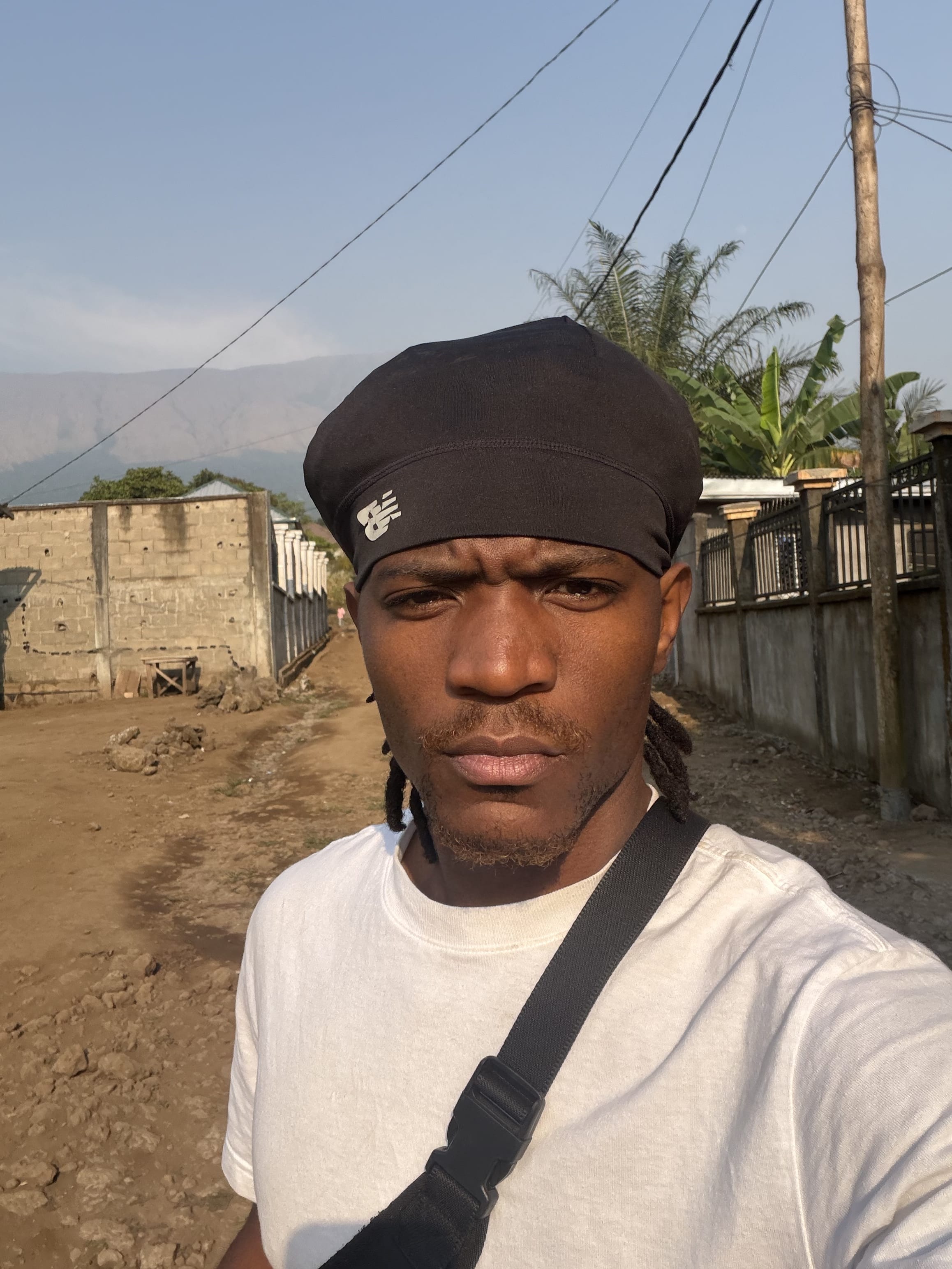 Selfie in a black durag on a red-dirt road - breeze-block walls, palms, and mountain haze behind