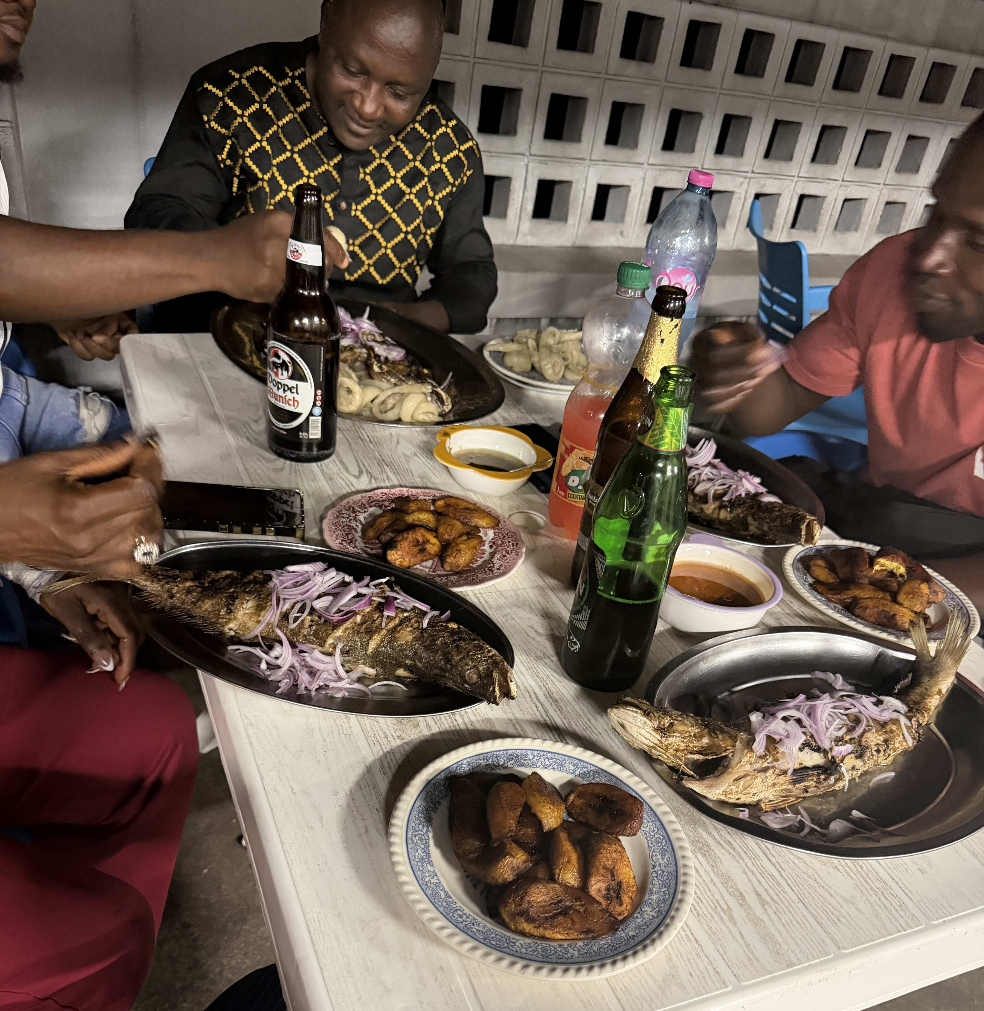 A table crowded with grilled fish under onions, fried plantains, and bottles - dinner with the family