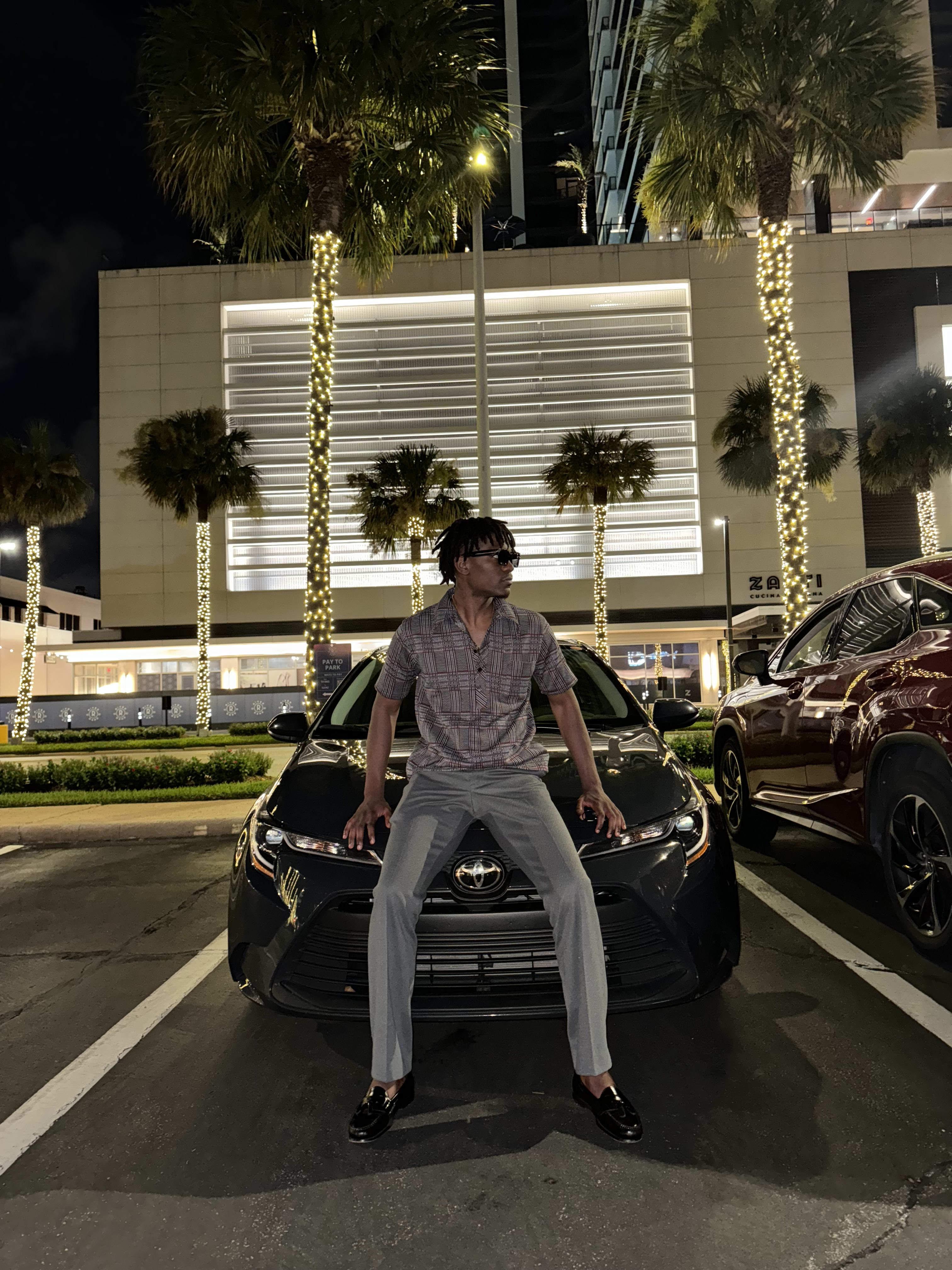 Kama seated on the hood of a black Toyota at night between palm trees wrapped in lights
