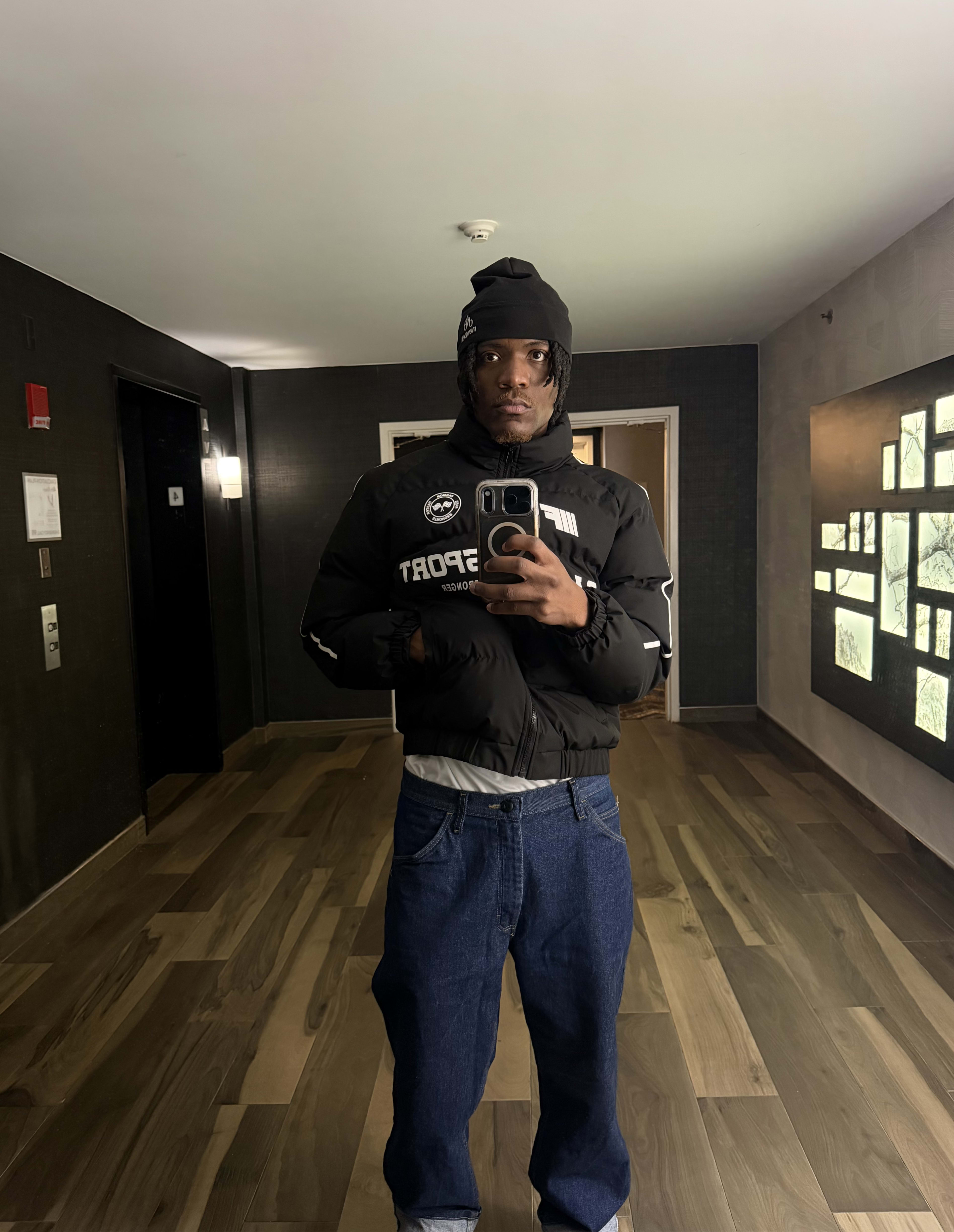 Mirror selfie in a black beanie, Sport puffer, and baggy jeans in a wood-floored corridor