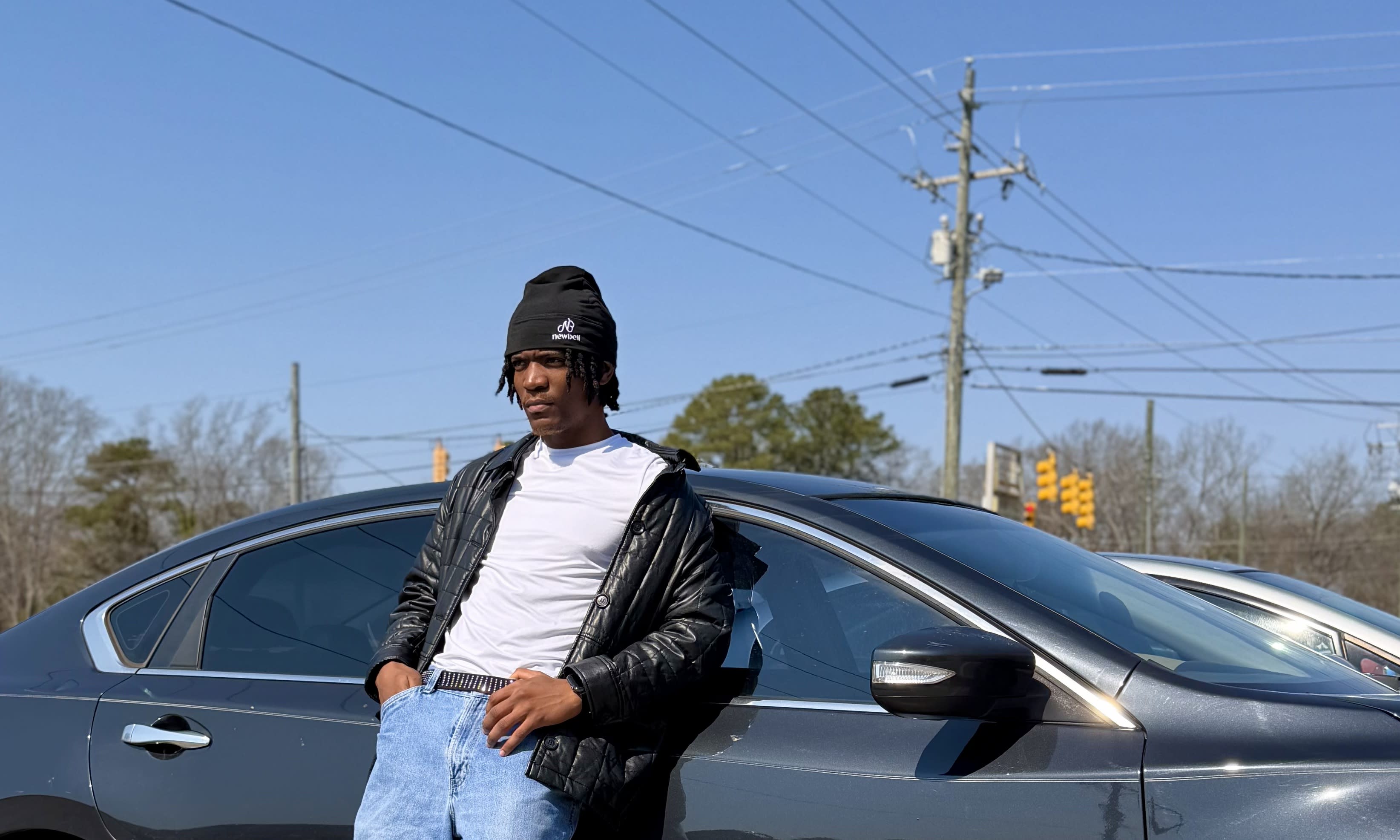 Kama leaning on a sedan in a black puffer and beanie, roadside power lines behind