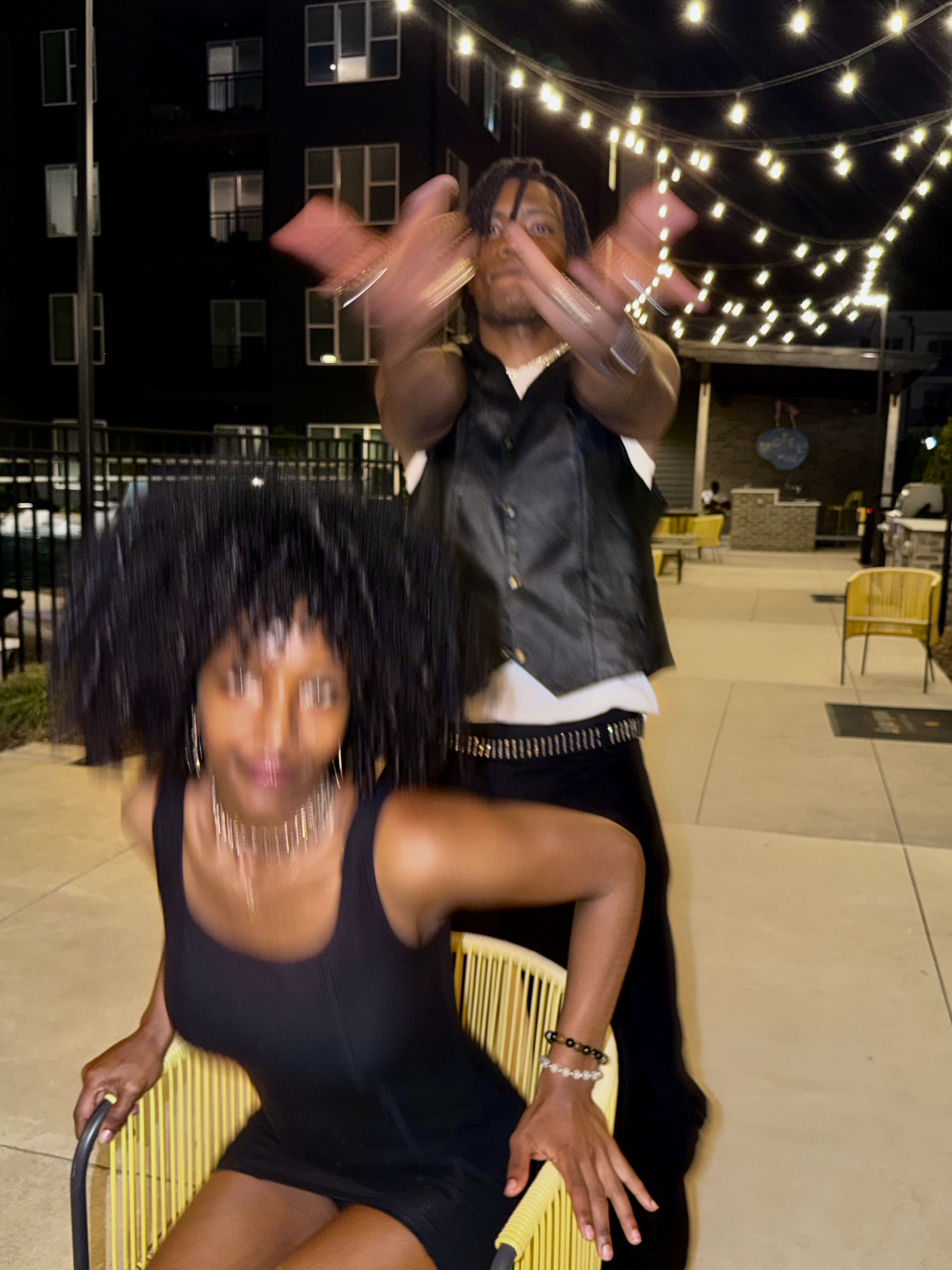 Motion blur under patio string lights - Kama in a leather vest dancing behind a friend on a yellow chair