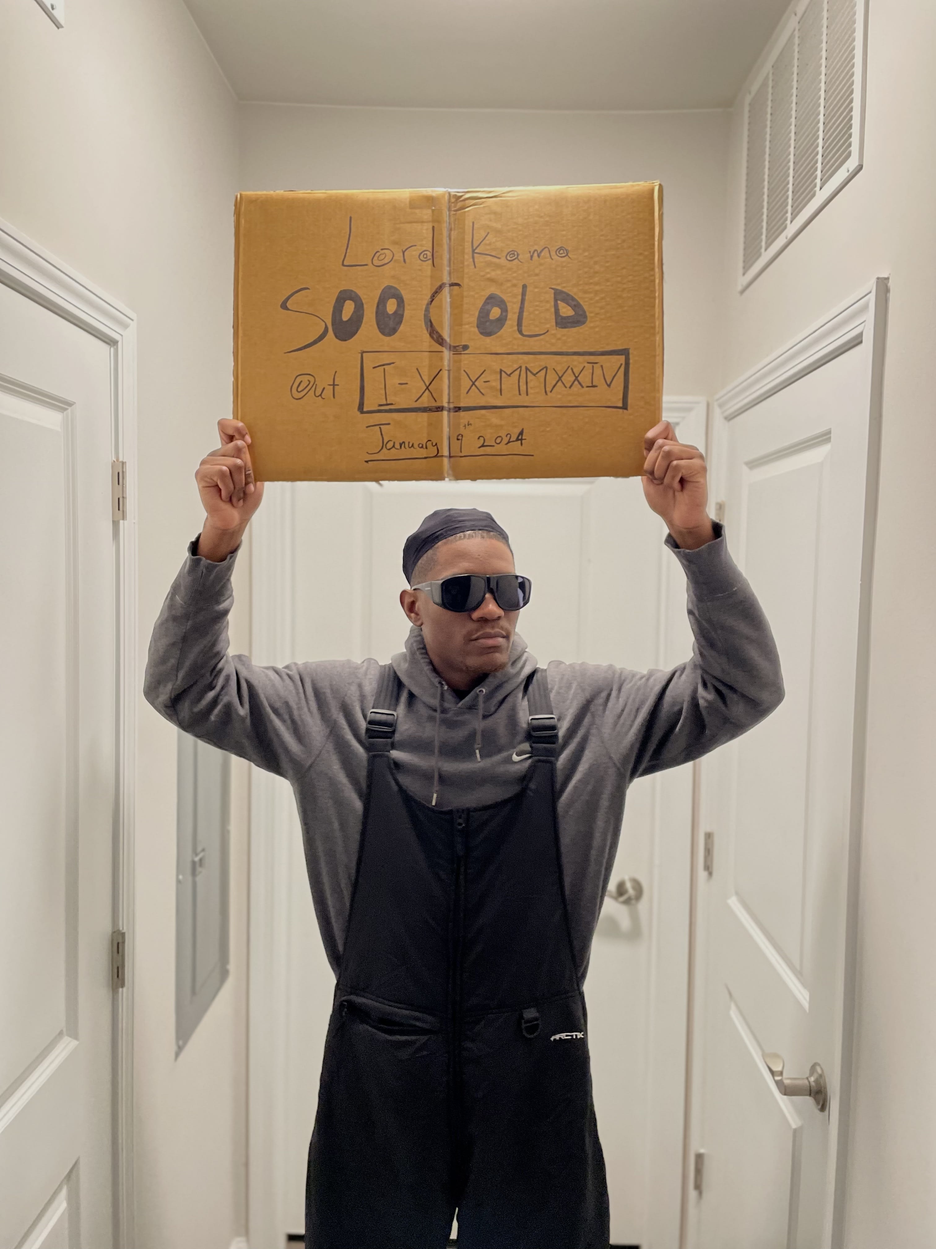 Kama in shades and overalls holding a cardboard sign overhead: Lord Kama, Soo Cold, January 19 2024