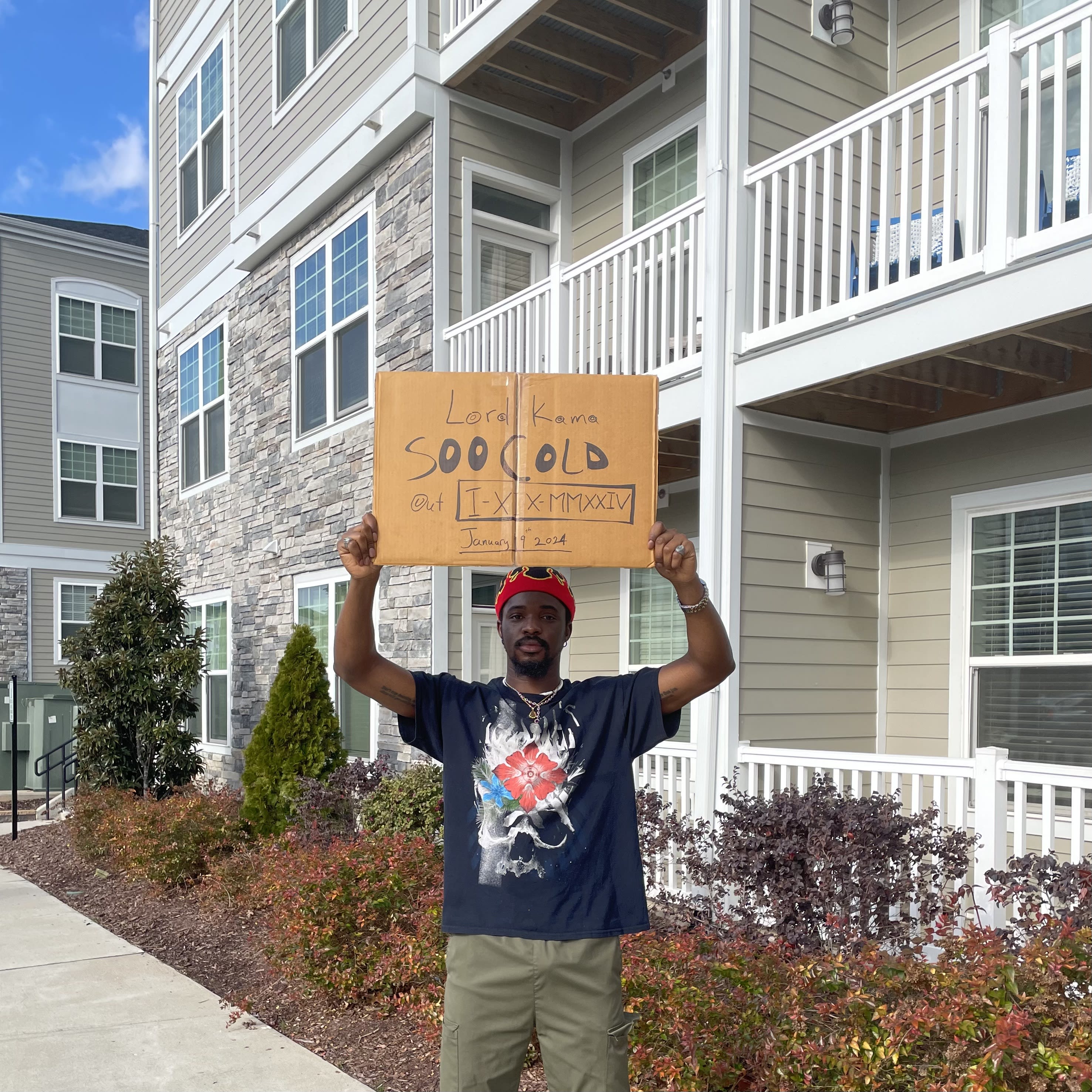 Kama outside the apartments holding the cardboard Soo Cold release sign overhead, red beanie on