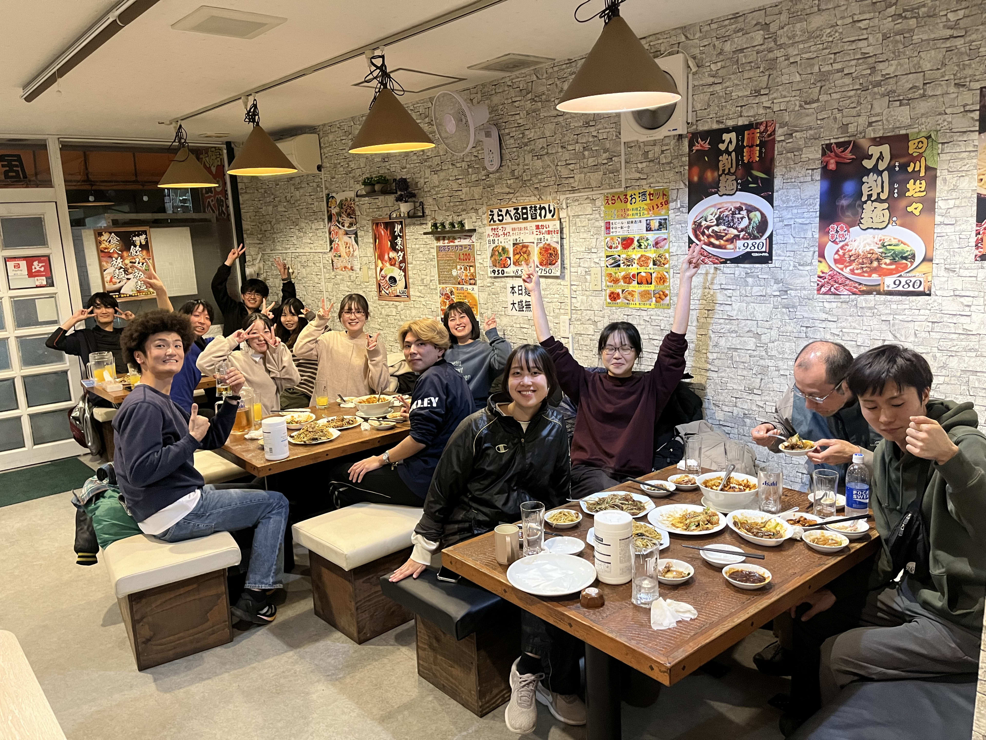 Friends waving over plates at a noodle restaurant in Japan, menus papering the walls