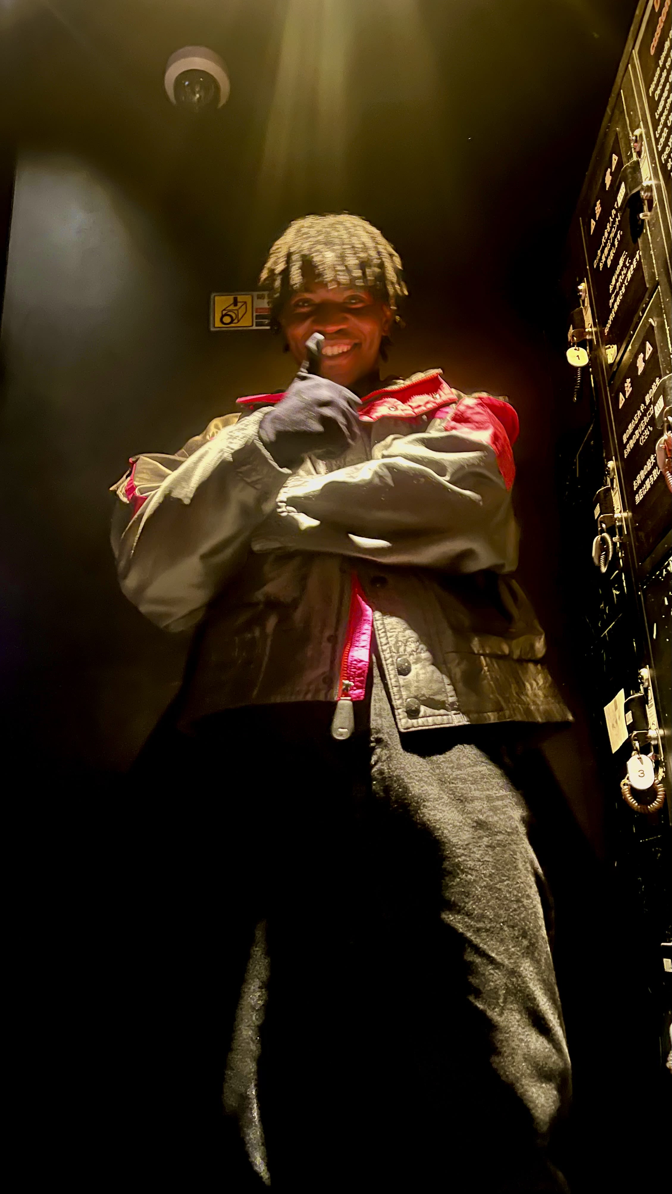 Low-angle grin in an elevator - frosted curls, gloves, silver-and-red racing jacket