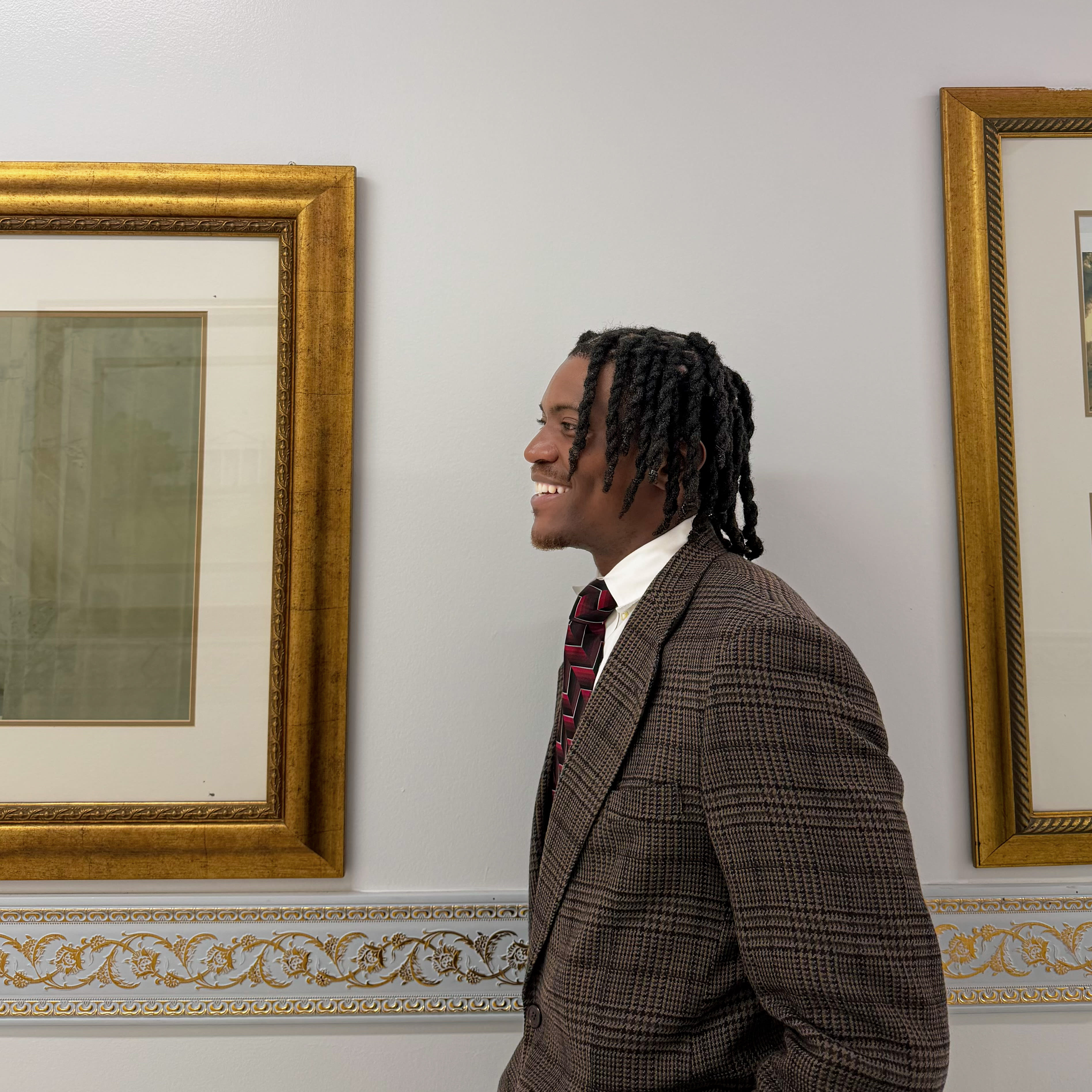 Kama laughing in profile - brown plaid blazer, striped tie, braids, gilded frames on the wall