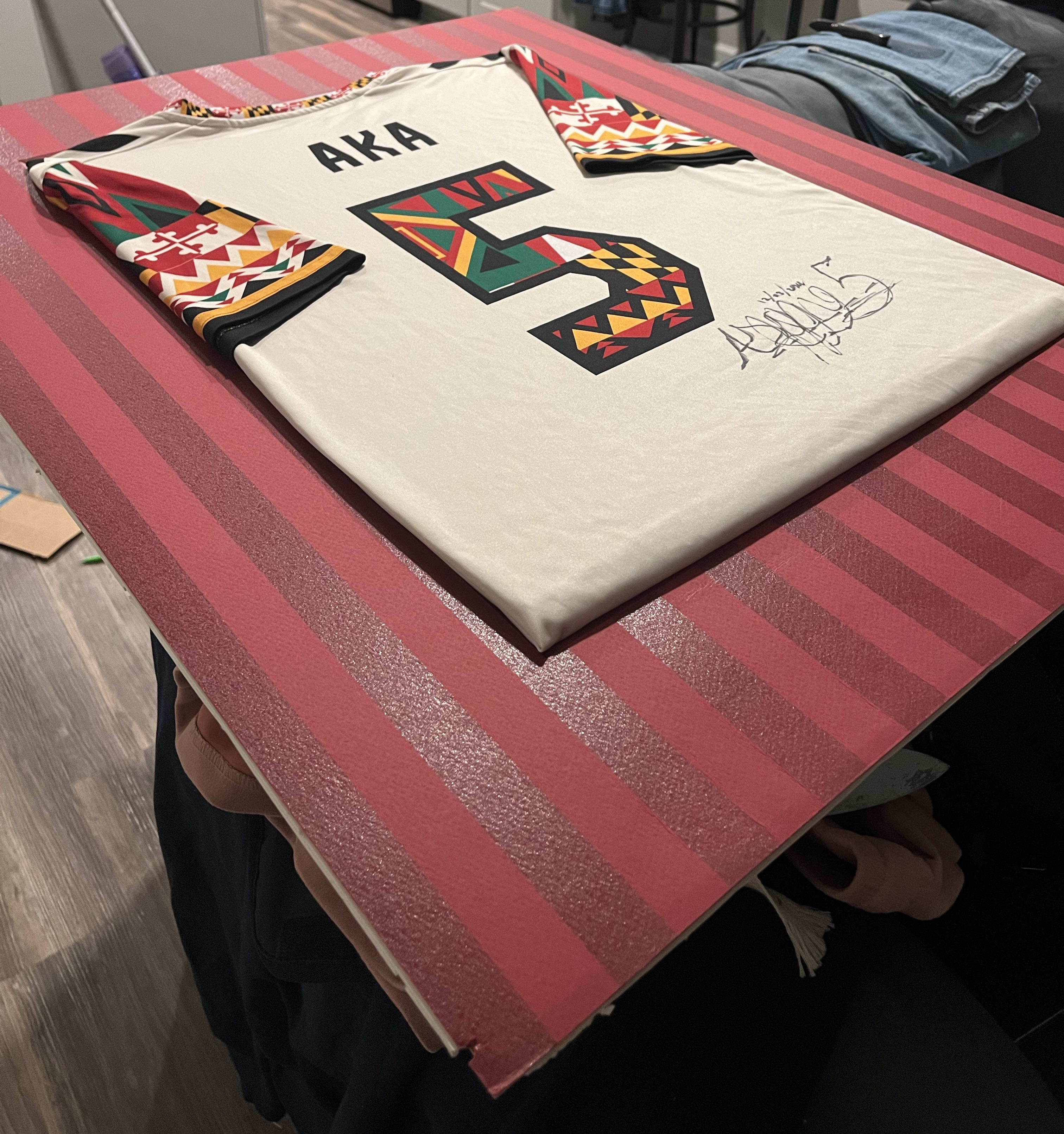 A framed white AKA 5 football jersey with African-print trim, signed across the chest