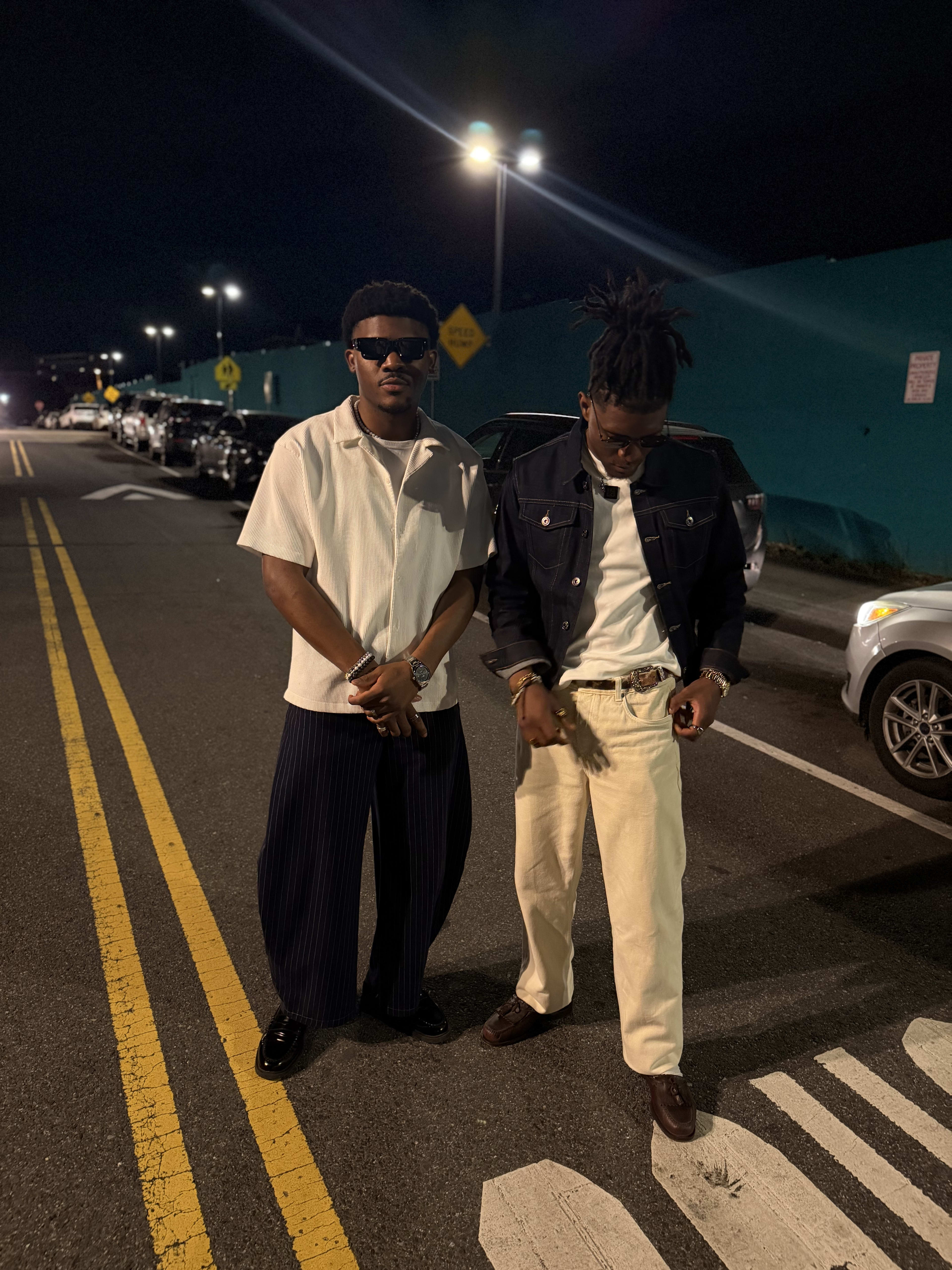 Kama in a white camp shirt and pinstripes with a friend in denim, night street behind them