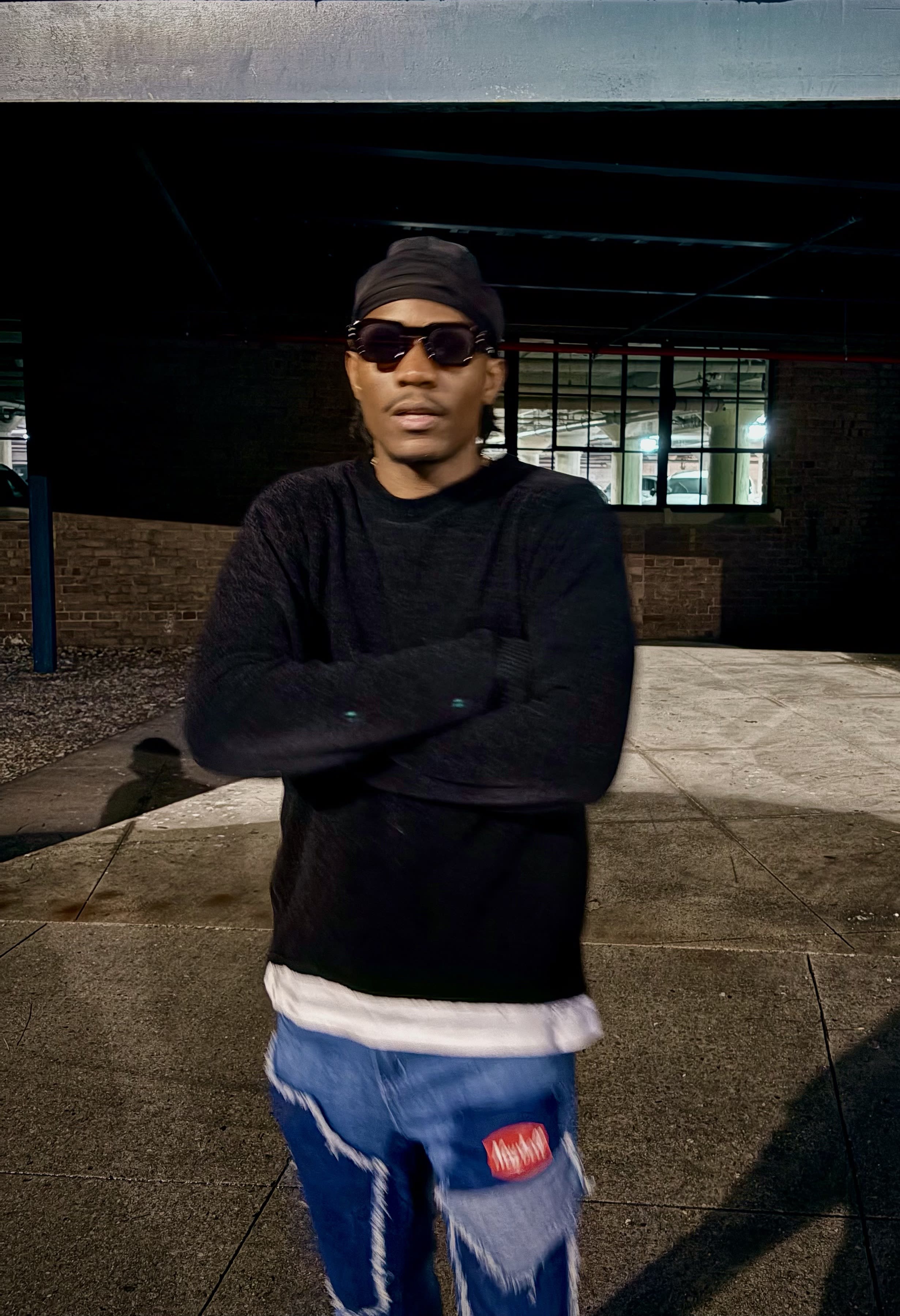 Kama in a black fuzzy sweater, shades, and head-wrap, arms crossed in a night parking deck