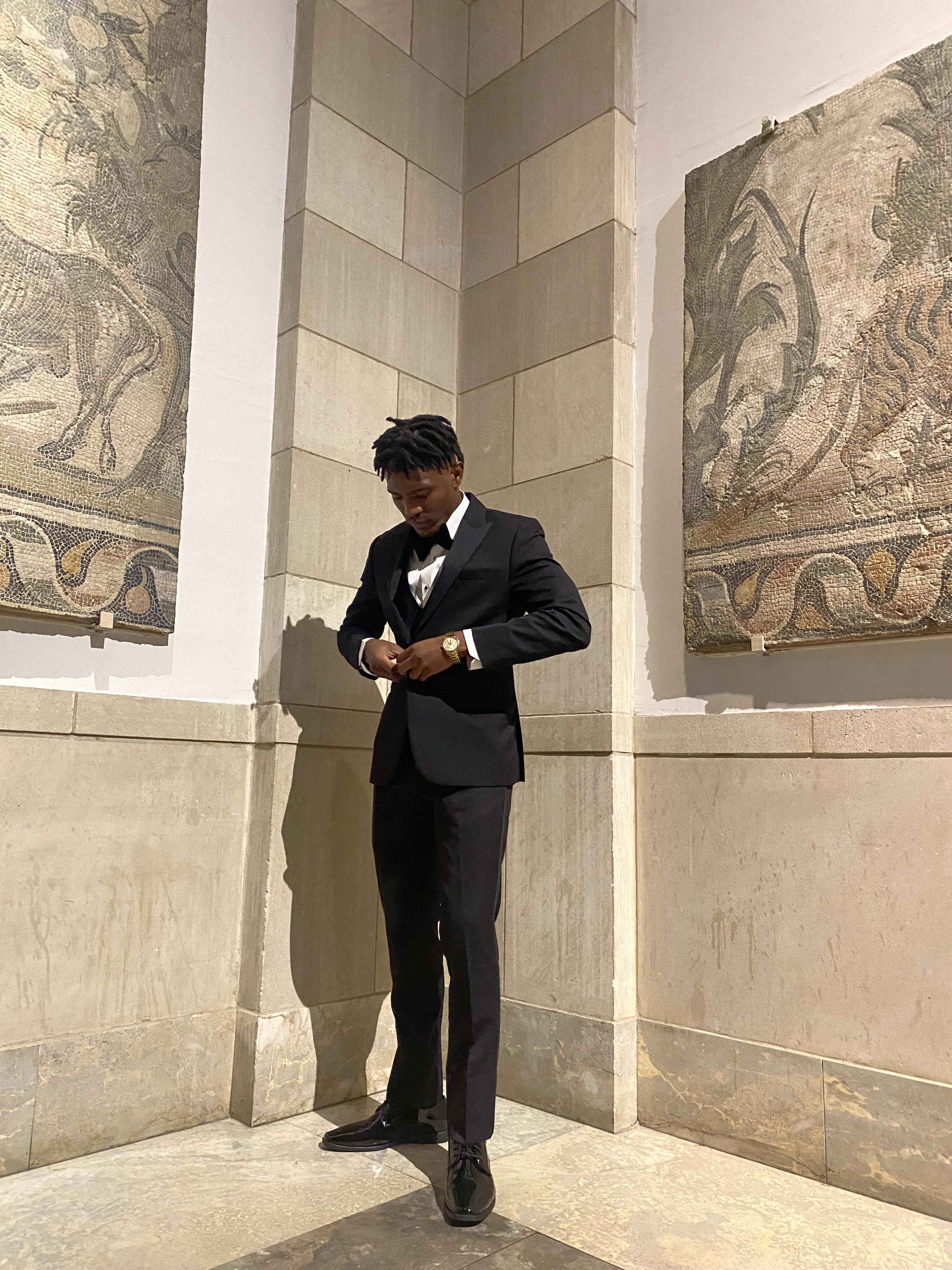 Kama in a black tuxedo and gold watch between ancient mosaic panels at the museum